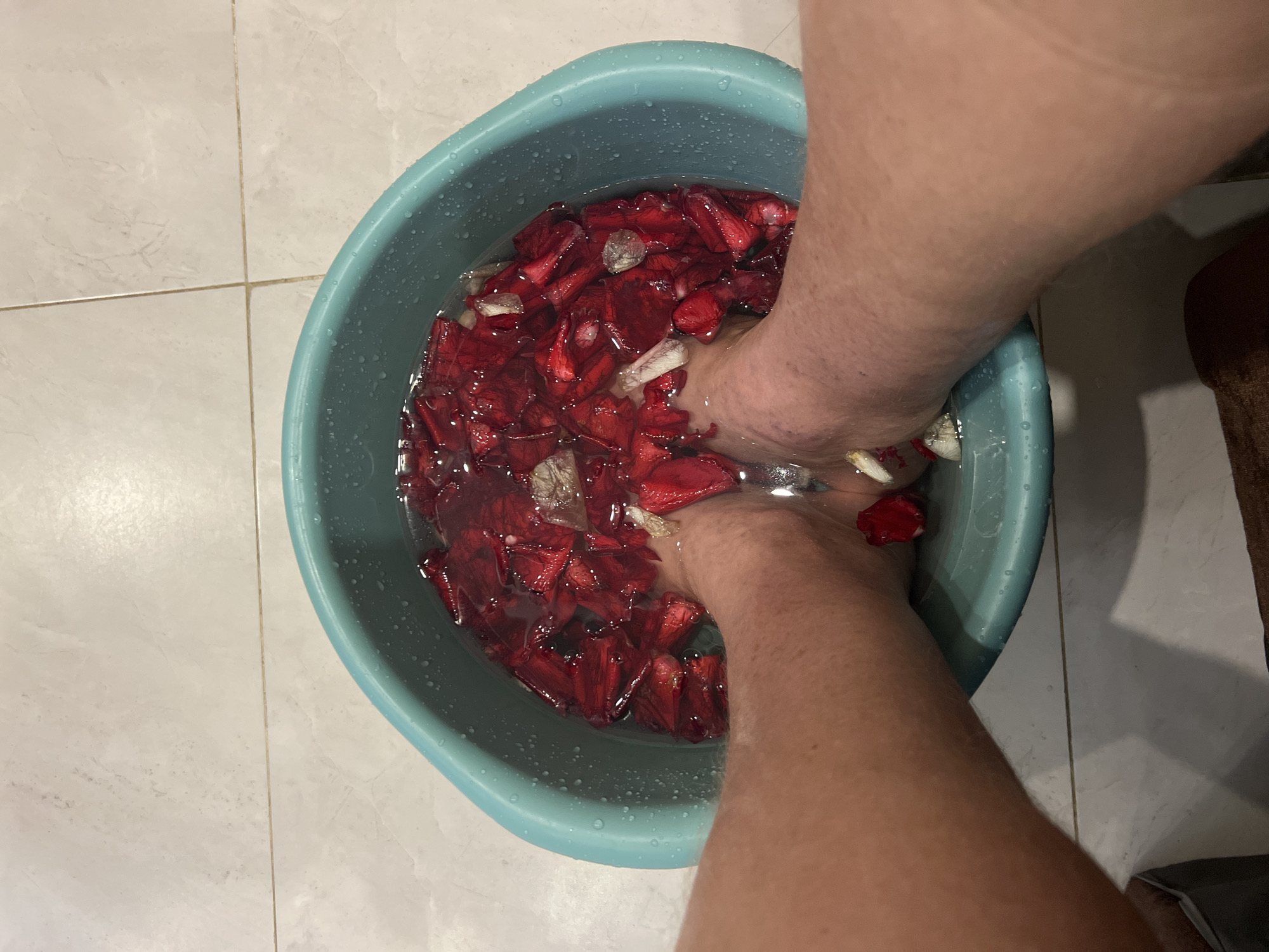 The image captures a top-down view of a person's feet soaking in a blue-green plastic basin filled with water and numerous red petals, along with a few lighter-colored petals or plant fragments. The setting is indoors, likely in a bathroom or spa area, indicated by the light-colored tiled floor with visible grout lines.
The primary subjects are two bare feet and lower legs, partially immersed in the water. The activity depicted is a foot bath or foot soak, possibly for relaxation or a therapeutic purpose, given the presence of the petals, which suggest a natural or aromatic treatment.
Notable details include the vibrant red petals, which are abundant in the water and surround the feet. The water appears still and clear, reflecting a slight sheen. The blue-green basin is simple and utilitarian, with water droplets visible on its exterior rim. The tiled floor beneath the basin is clean and well-maintained. There is no visible text in the image. The high-angle perspective places the viewer directly above the activity. The location context provided is Bac Nha Trang Ward, Vietnam, suggesting this foot bath is taking place in that region.