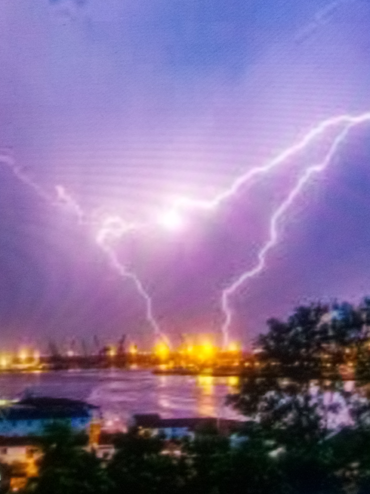 This image captures a dramatic lightning storm over a harbor at dusk. The sky is a deep purple, illuminated by several bright bolts of lightning that stretch across the frame. In the foreground, a body of water reflects the city lights and the ominous sky. Along the waterfront, there are industrial structures, possibly cranes and buildings, with numerous yellow lights twinkling. The foreground also shows the tops of trees and several buildings, one of which is a light blue and white structure. The overall atmosphere is intense and awe-inspiring, with the raw power of nature on full display over a city landscape.