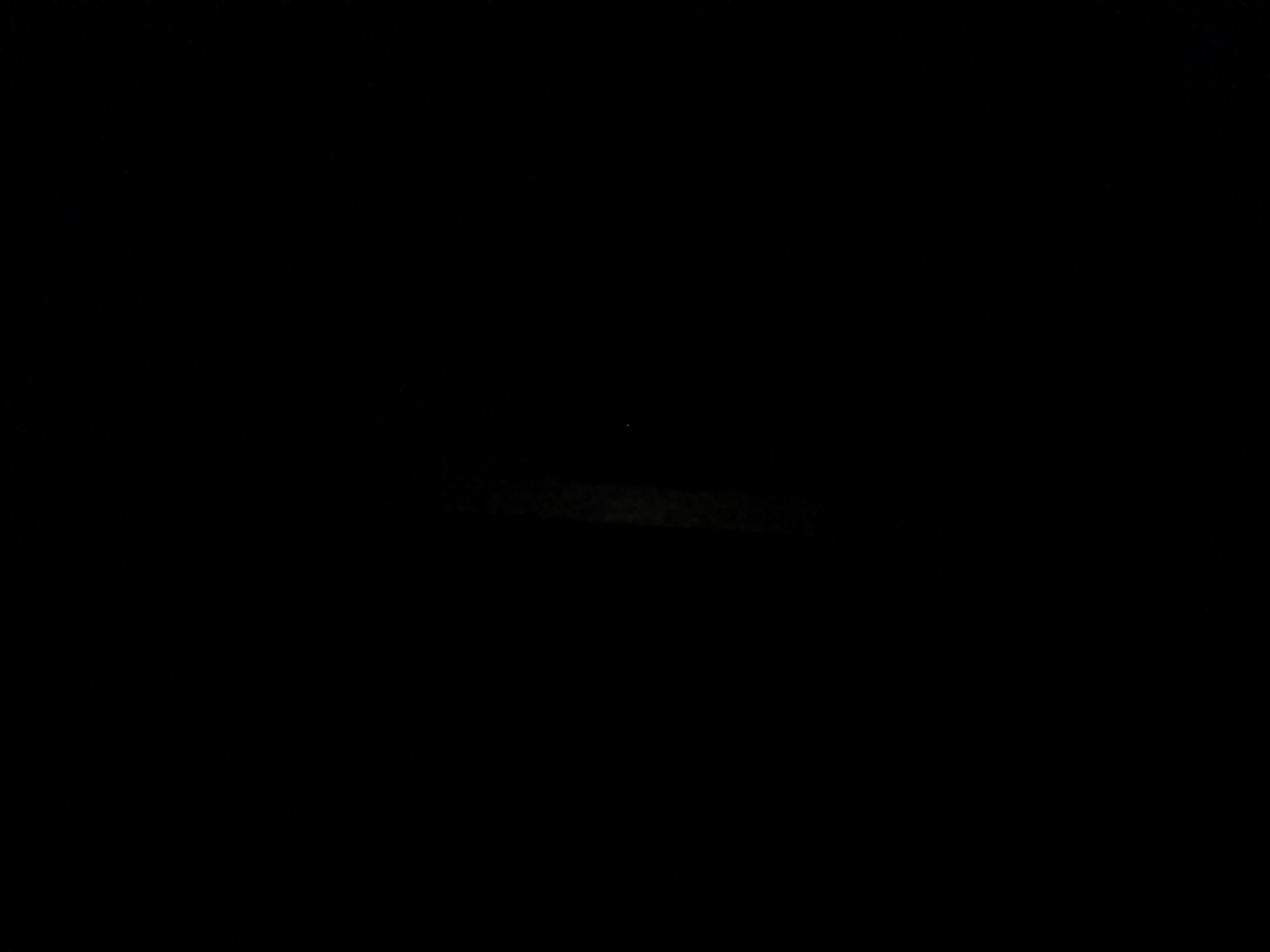 This image shows a vertical, slightly out-of-focus view of a dimly lit interior space, likely a building in Kaunas, Lithuania. The primary subject is a vertical shaft of light that appears to be emanating from a window or opening high up on the left side of the frame. This light illuminates a portion of a dark, textured wall, revealing faint vertical lines or a subtle pattern. The rest of the image is predominantly black, suggesting a lack of ambient light and making it difficult to discern any other details of the environment. No people, objects, or specific architectural features are clearly identifiable beyond the implied wall and the light source. The overall impression is one of an empty, unlit space with a single source of illumination.