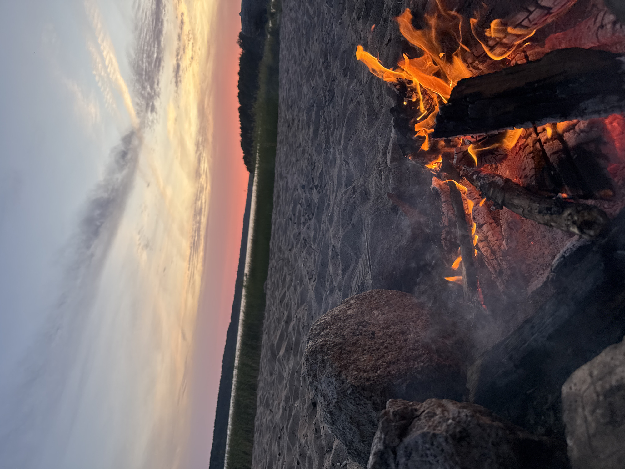 The image depicts a beach scene at dusk with a bonfire. The foreground shows a crackling bonfire with burning logs and embers, surrounded by sand and a few large rocks. In the mid-ground, the sandy beach extends towards a body of water, beyond which a tree-lined shore is visible. The background features a colorful sky with hues of pink, orange, and blue, indicating sunset. There is no indication of the city of Красная Долина, Russia, in the image.