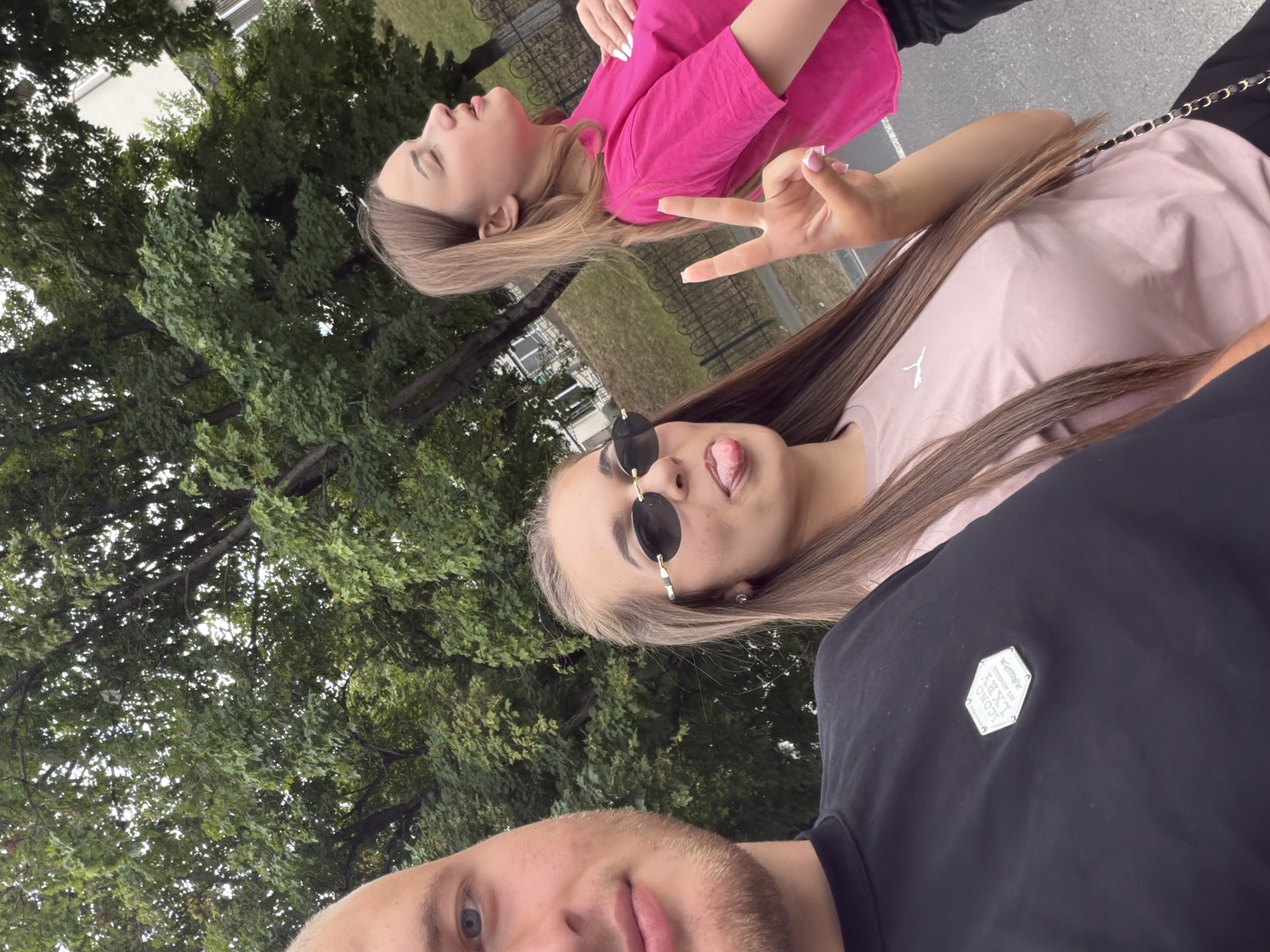 In Strășeni, Moldova, a selfie captures three individuals outdoors, with lush green trees forming the background. In the foreground on the left, a man with light-colored hair and blue eyes is partially visible, wearing a black t-shirt with a hexagonal emblem. To his right, a woman with long brown hair and sunglasses is sticking out her tongue and giving a peace sign. She is wearing a light pink t-shirt. Further to the right, another woman with blonde hair is wearing a bright pink t-shirt and looking upwards. The scene appears to be a casual outdoor setting, possibly a park or a street, with a metal fence and buildings visible in the distance. The lighting suggests it is daytime.