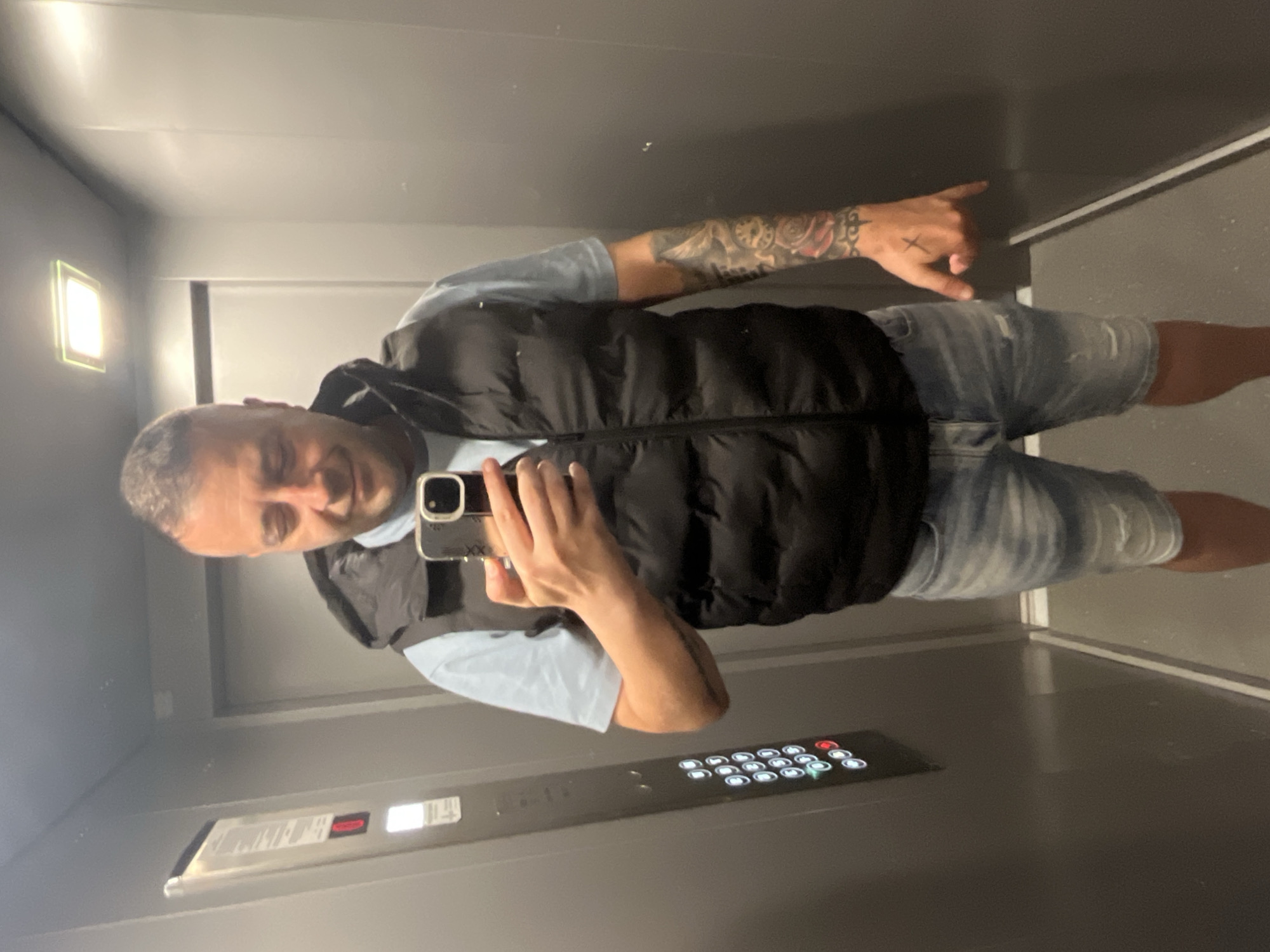 A man is standing in an elevator, taking a selfie with his phone. He is wearing a light blue t-shirt, a black puffer vest, and distressed denim shorts. He has tattoos on his left arm and a small cross tattoo on the back of his right hand. The elevator has grey walls, a control panel with buttons, and a light fixture on the ceiling. The image is taken from a slightly low angle, looking up at the man. The scene does not contain any elements that would allow for determining a specific location in the city of Varna, Bulgaria.