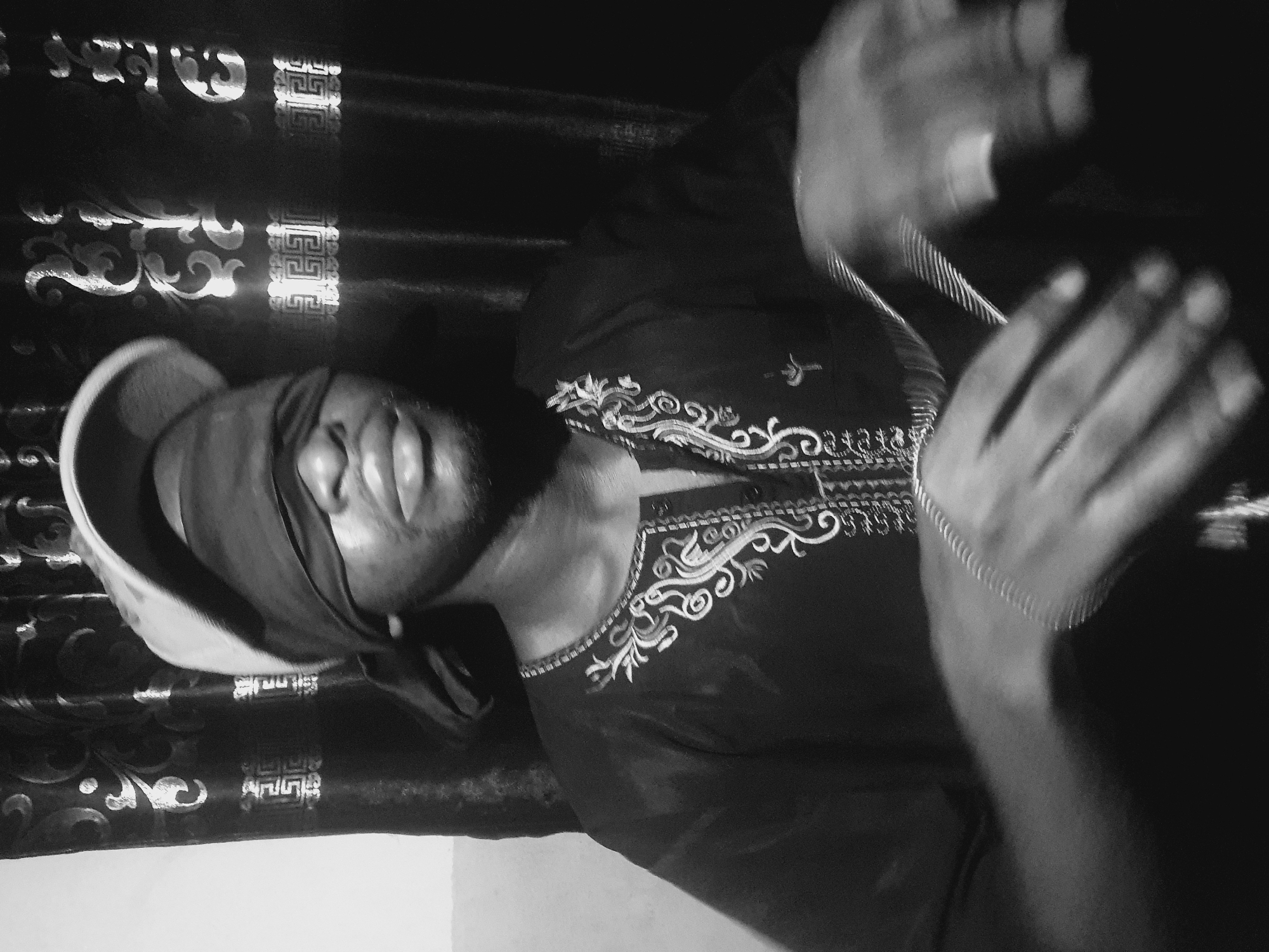 A monochrome image depicts a man indoors, possibly in Lokoja, Nigeria. He is blindfolded with a dark cloth and wears a light-colored cap. He also wears a dark garment with intricate, light-colored embroidery around the neckline. His hands are raised and slightly blurred, suggesting movement, and a silver bracelet is visible on his wrist. The background includes a patterned dark curtain or fabric with ornate designs. The base of a white wall is also visible. The orientation of the image is horizontal, with the subject's head tilted to the left. The scene has a dramatic, high-contrast lighting.