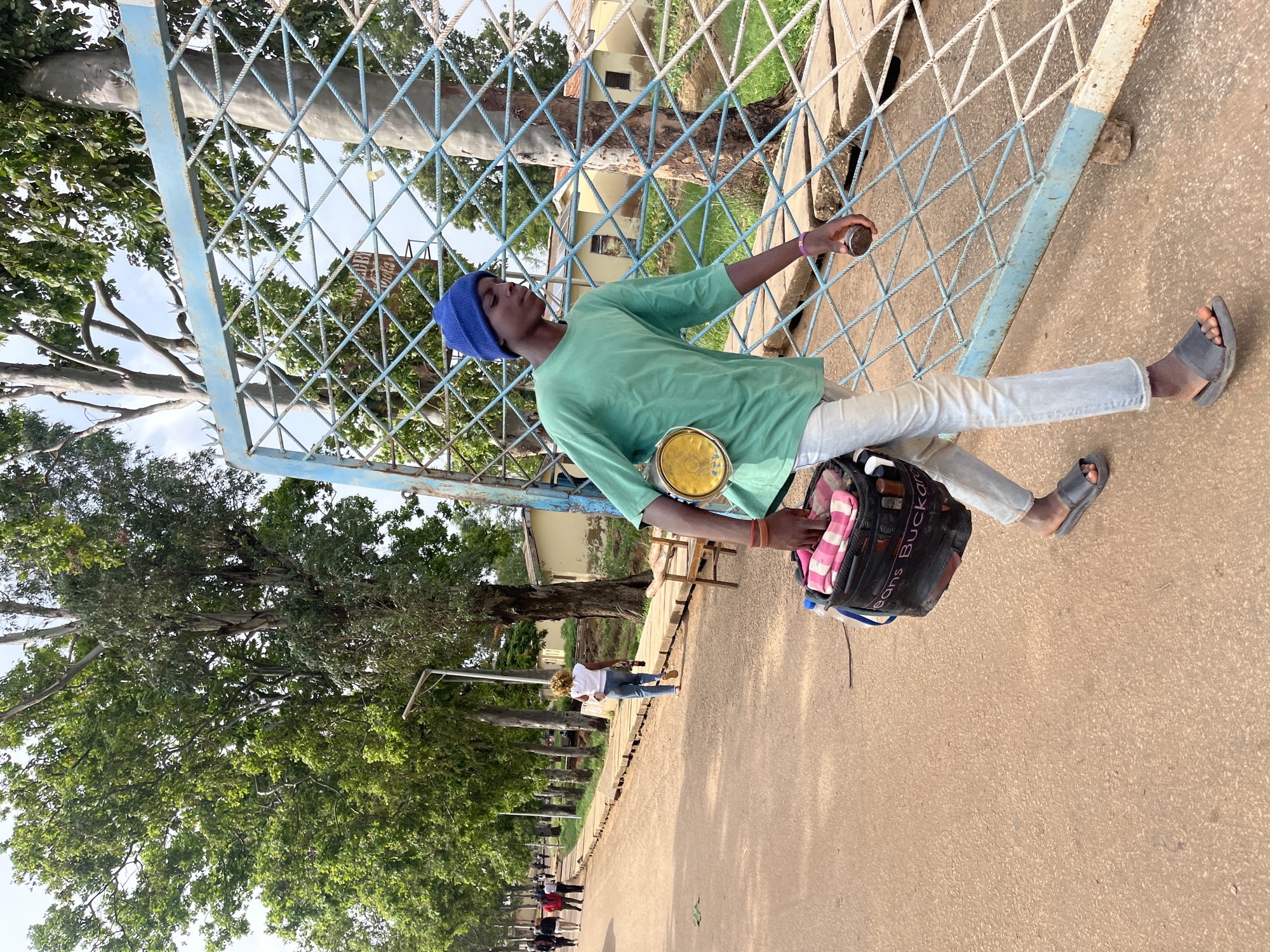 A young man walks past a blue metal gate in Rafin Par, Nigeria. He wears a green t-shirt, a blue beanie, and light-wash jeans, and carries a black bucket with pink and white fabric peeking out. He holds a small, round object in his right hand. The gate is part of a fence that runs along a dirt path lined with trees. In the background, other people walk along the path, and buildings are visible. The overall environment appears to be an outdoor urban setting.