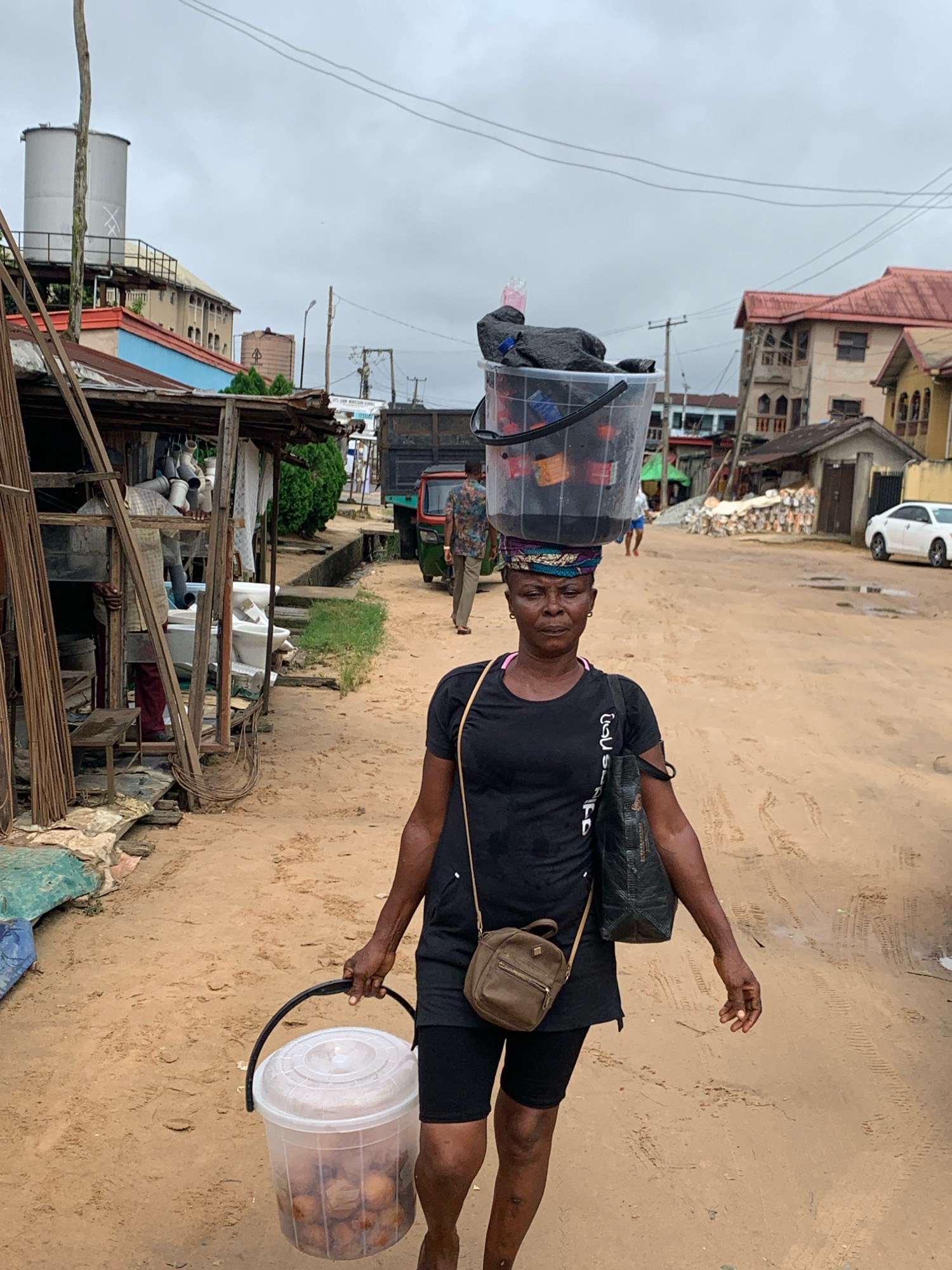 A woman is walking down a dirt road in Umuode, Nigeria, carrying a large, clear plastic bucket filled with items on her head, balanced by a black plastic bag and a dark-colored fabric item stuffed into it. She is also carrying another clear plastic bucket by its handle in her right hand, which contains round, light-brown objects. She is wearing a black t-shirt with white lettering, black shorts, and a patterned headwrap. A small brown purse is slung across her body. In the background, several buildings with red roofs are visible, along with a white car parked on the side of the road. To the left of the woman, there is a stall with various items displayed, including what appear to be pipes and sanitary ware. Power lines crisscross the sky, and the ground is a dusty, sandy color with some tire tracks. The overall environment suggests a bustling, outdoor market or street scene in a Nigerian town.