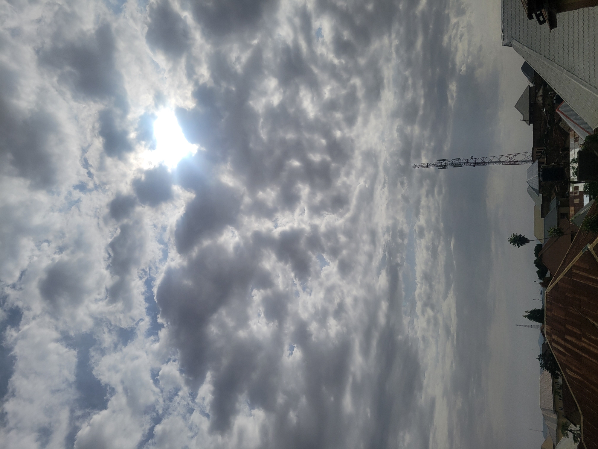 The image displays a daytime scene over Jos, Nigeria, dominated by a dynamic sky. The sky is largely covered by a mixture of dense, dark grey clouds and brighter white clouds, with patches of blue sky visible in between. The sun, a brilliant white orb, is prominently featured in the upper left quadrant, partially obscured by clouds but shining intensely, creating a strong glare and visible rays of light.
Below the expansive sky, a distant urban or semi-urban landscape stretches across the horizon. A tall, slender metal lattice telecommunications tower stands out against the cloud-filled sky, positioned slightly to the right of the center. Various buildings and rooftops are visible, suggesting a populated area. These include structures with light-colored, possibly corrugated metal roofs, and others with darker finishes. In the foreground on the right, a reddish-brown, likely rusty, corrugated iron roof is discernible. Scattered among the buildings are a few trees, including a distinct palm tree to the right of the tower.
The time of day is clearly daytime, indicated by the high and intense sun. The weather is partly cloudy to overcast, with strong sunlight piercing through the cloud cover. There is no visible text, and no explicit human activity or events are captured in the static landscape and sky.