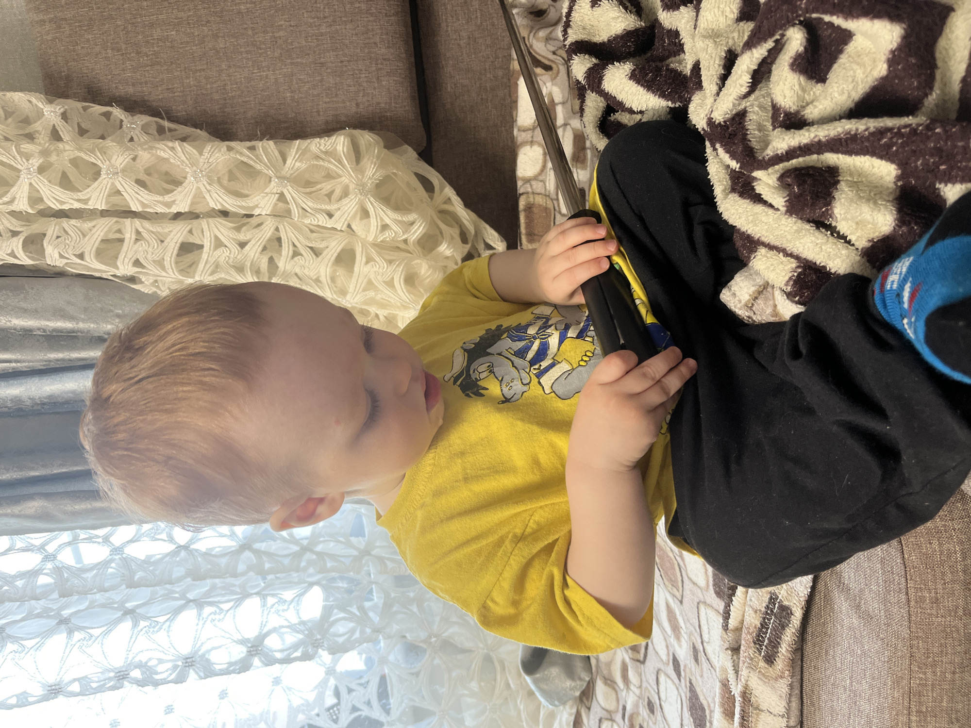 A young child sits on a brown couch in an indoor environment. The child is wearing a yellow T-shirt with a cartoon design and black pants. A blanket with a brown and white swirling pattern covers the lower half of their body, with a blue sock visible on their right foot. The child is holding a black object, possibly a remote or electronic device, in their hands. Beside the child is a section of white lace fabric, and the couch has a textured brown surface. The location is indicated as Chaikovsky, Russia.