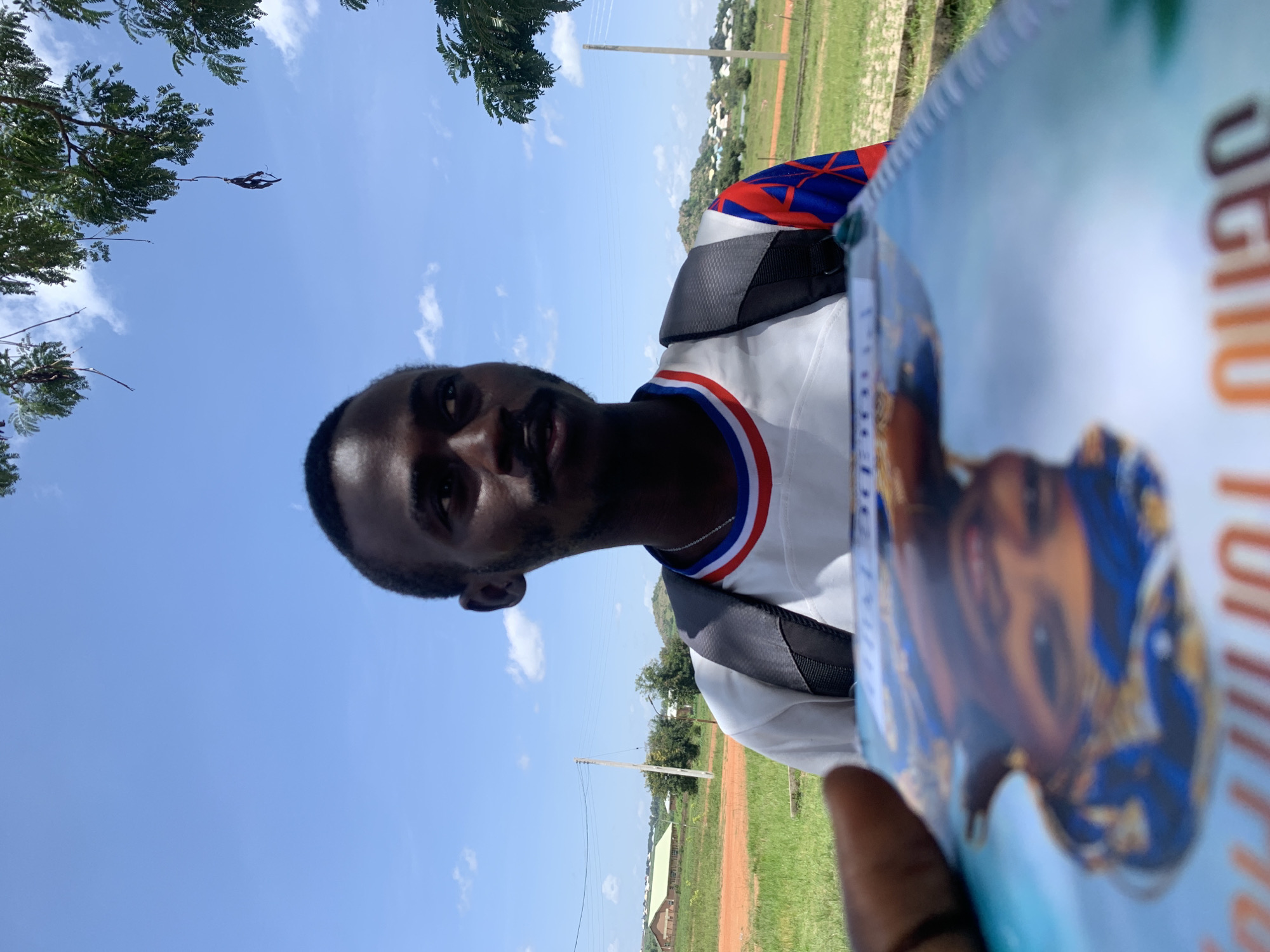 A close-up, low-angle, over-the-shoulder shot shows a young Black man looking to his right, with a book held in the foreground, obscuring the lower portion of the image. The man is wearing a white t-shirt with red, white, and blue trim around the collar and a blue, red, and white patterned design on the right sleeve. He has a short beard and mustache. A dark backpack with grey straps is visible on his shoulders. The background consists of a bright blue sky with scattered white clouds, green grass, a dirt path, and some buildings and trees in the distance. The presence of a utility pole with wires and the architecture of the distant buildings suggest an outdoor, possibly rural or semi-rural, setting within Nigeria, specifically Gwafan, which is noted for its open spaces and scattered settlements. The book in the foreground has an illustration of a person's face and some text, but it is blurred and difficult to decipher. The lighting indicates a sunny day.