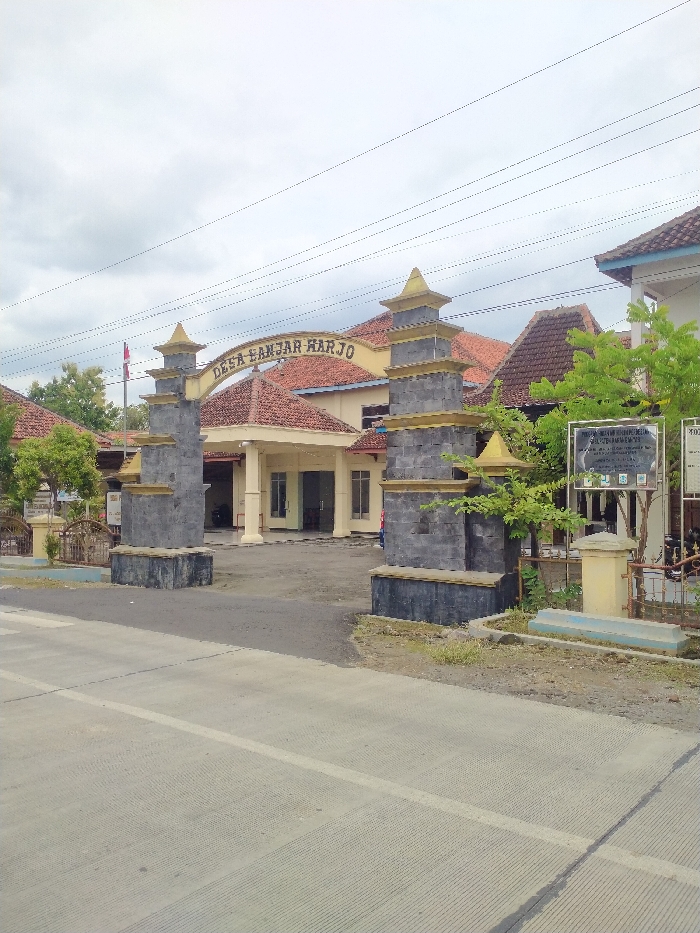 The image presents a view of the entrance to Desa Banjar Harjo (Banjar Harjo Village) in Kecamatan Kebakkramat, Indonesia. A prominent gateway, composed of two substantial pillars made of grey stone blocks and topped with golden-yellow pyramid-like caps, frames the access point. An arched sign spanning between the pillars clearly displays the text "DESA BANJAR HARJO" in golden-yellow lettering.
Behind the archway, a single-story building with light-colored walls and a reddish-brown tiled roof, likely a village office or community center, is visible. The architecture features multiple gables and a covered porch area. Green trees and shrubs are interspersed around the entrance and alongside the buildings, contributing natural elements to the setting. On the right side of the archway, two informational signs are positioned; one partially legible sign begins with "PEMERINTAH KABUPATEN..." (Government of Regency...). A flag pole bearing a red and white flag, presumably the Indonesian flag, is visible further back on the left.
In the foreground, a grey concrete road with a subtle white line extends across the lower portion of the image. The sky is overcast, providing diffused lighting characteristic of a cloudy day. No people or specific activities are depicted, giving the scene a quiet and still atmosphere.