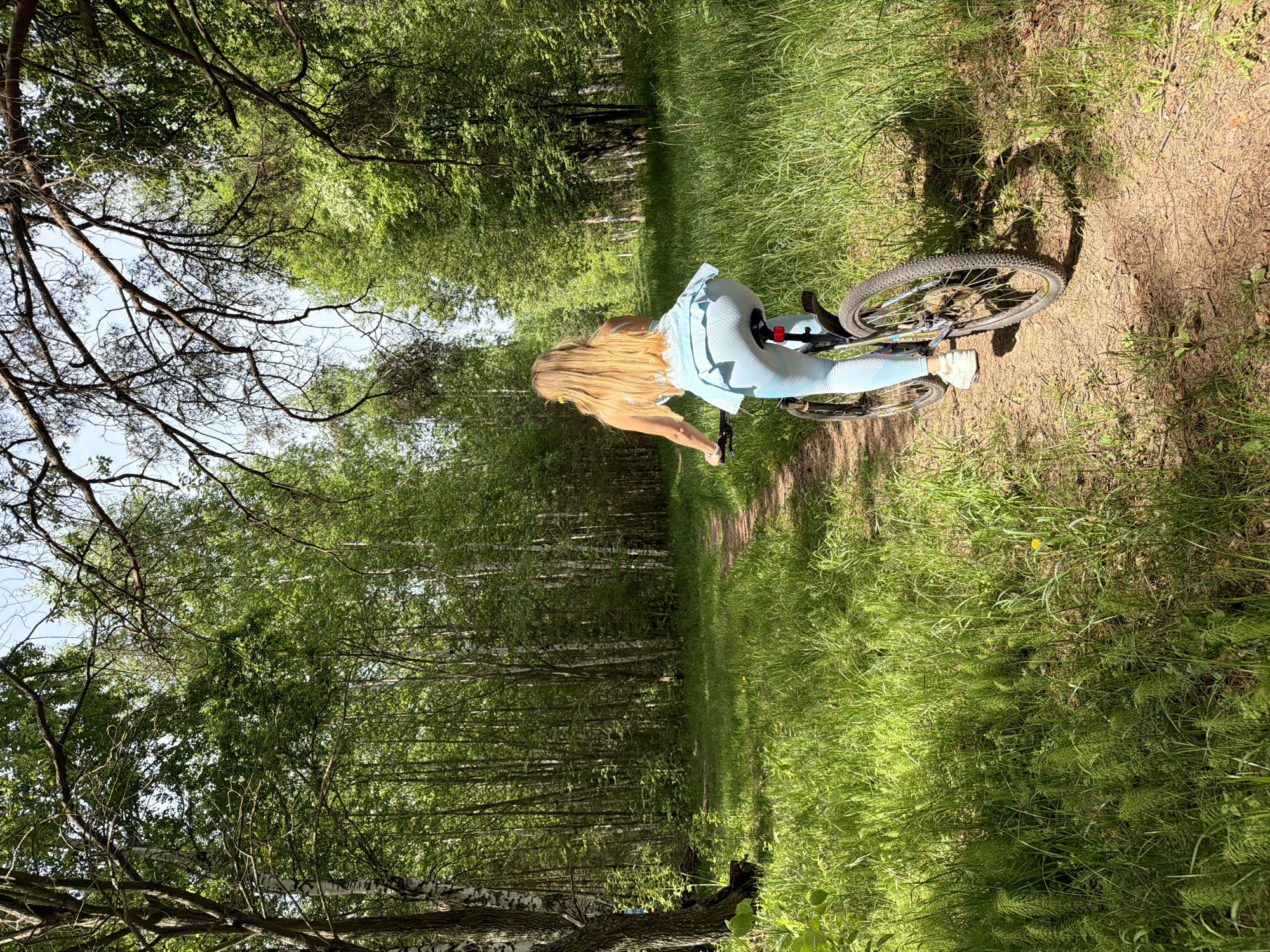 A blonde woman is riding a bicycle on a dirt path in a forest. The forest is in Chaikovsky, Russia, as indicated by the birch trees. The woman is wearing a light blue top and leggings. The path is surrounded by green grass and trees. The lighting suggests it is daytime.