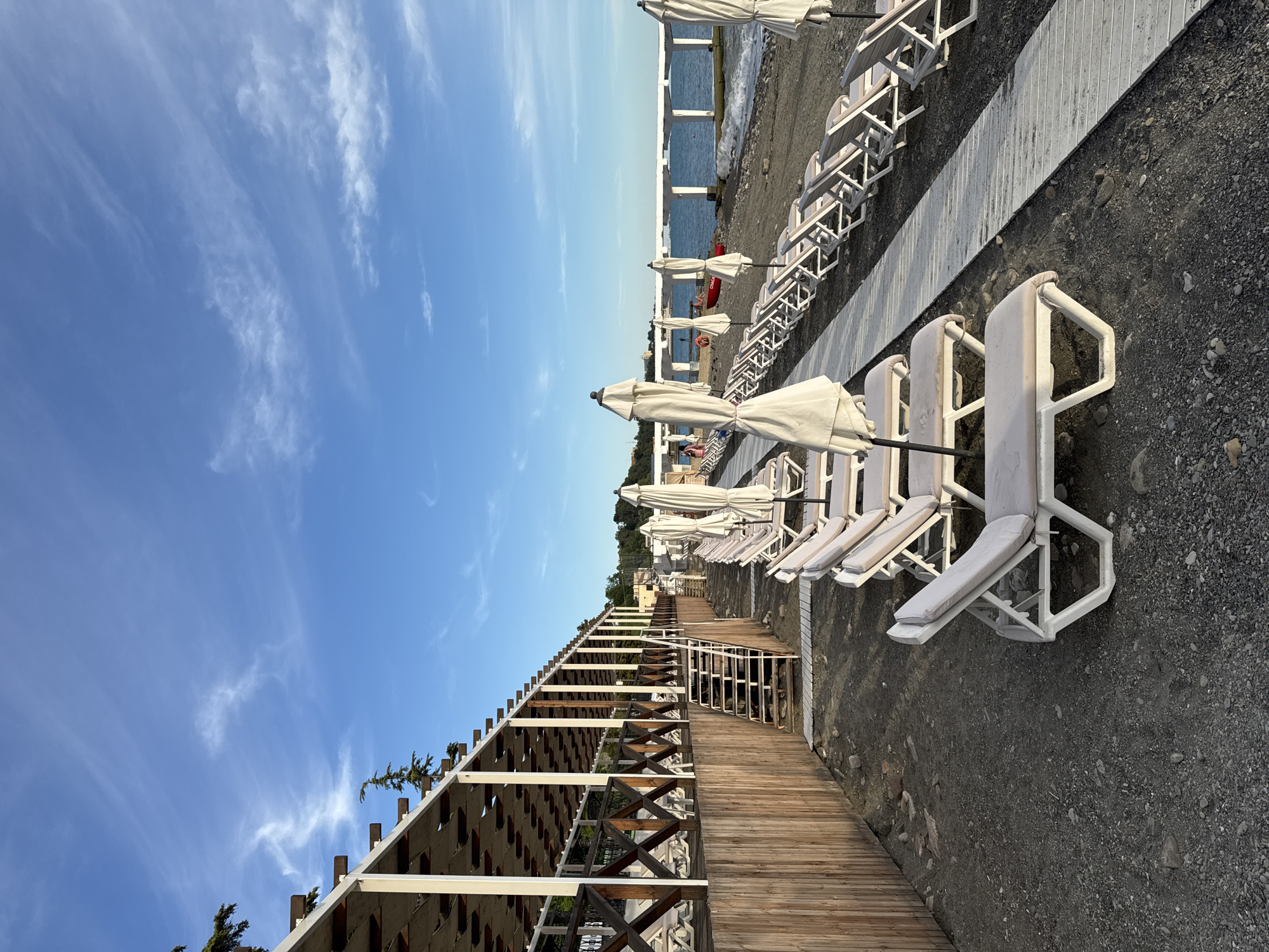A perspective shot captures an empty beach in Sochi, Russia, at dusk. Rows of empty lounge chairs and closed umbrellas line the dark pebble beach. A wooden boardwalk runs alongside the chairs, leading towards a pier in the distance. The sky is a brilliant blue with wisps of white clouds. A small red boat is visible near the pier. The scene suggests the end of a beach day, with the quiet atmosphere enhanced by the fading light.