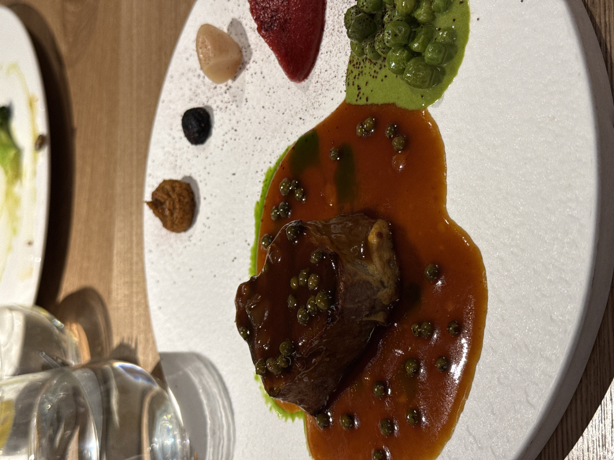 The image shows a plate of food, likely in a restaurant, with a piece of meat in a brown sauce as the central element. The meat is also covered with small, green round objects. Around the meat, there are various other components of the dish, including what appears to be a green purée with green peas, a red gel or purée, a pale beige blob, a small black ball, and a brown dollop. The food is arranged on a white, circular plate. The background includes a wooden table and partial view of glassware, indicating it's likely a restaurant setting. There is no information related to the location in the city of Krasnoyarsk, Russia.
