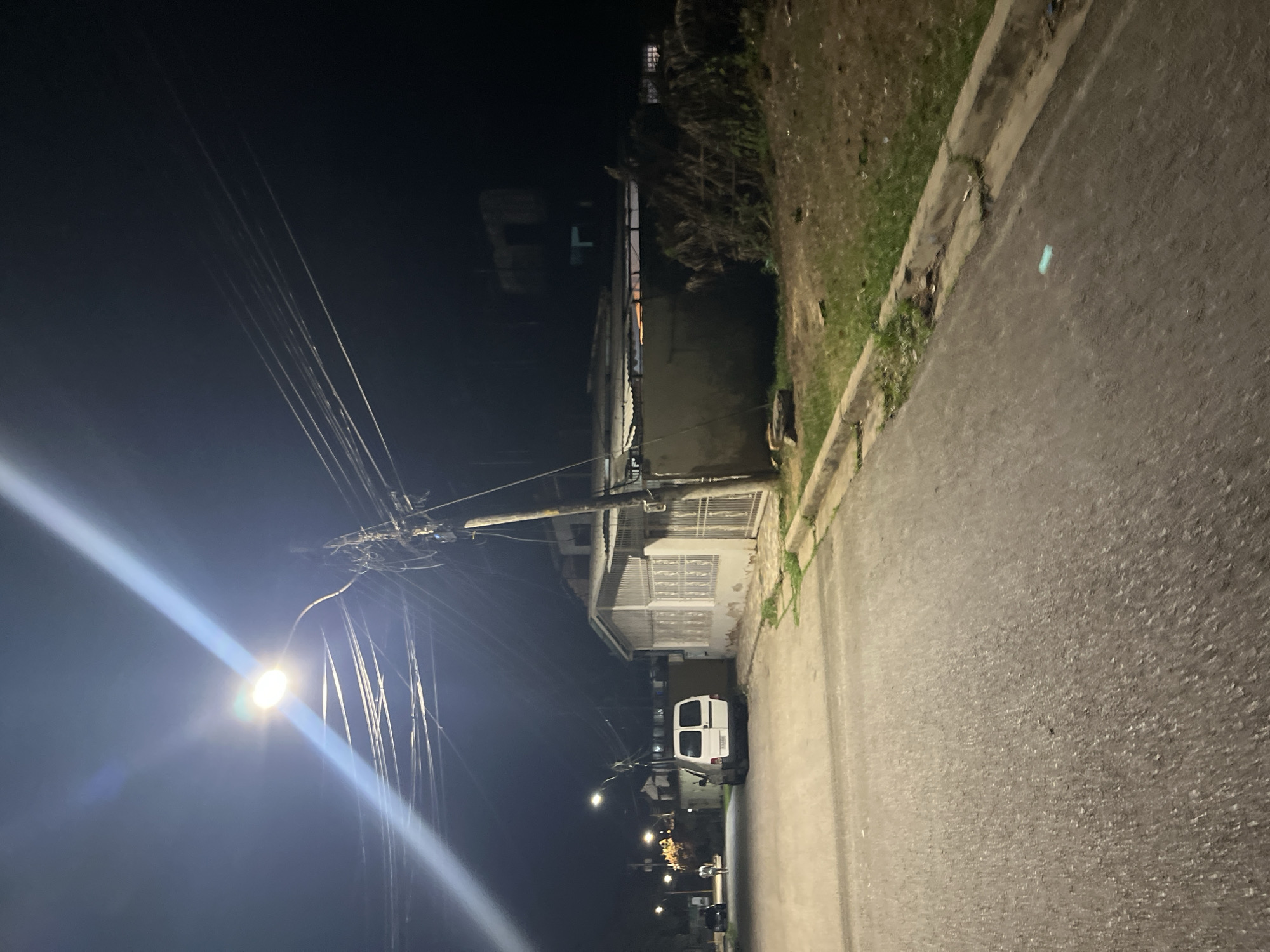 The image depicts a dimly lit residential street scene at night in Santa Ana, Venezuela. A white van is parked on the left side of the asphalt road, facing away from the camera. Further down the street, other vehicles and streetlights are visible, suggesting a somewhat populated area. A utility pole with numerous overhead wires stands prominently on the right side of the road, casting shadows and contributing to the overall cluttered overhead infrastructure. Adjacent to the pole, a house with a corrugated metal roof and white-paneled walls is visible. The area to the right of the road is a grassy verge with some dry foliage and debris. The sky is dark, indicating nighttime.