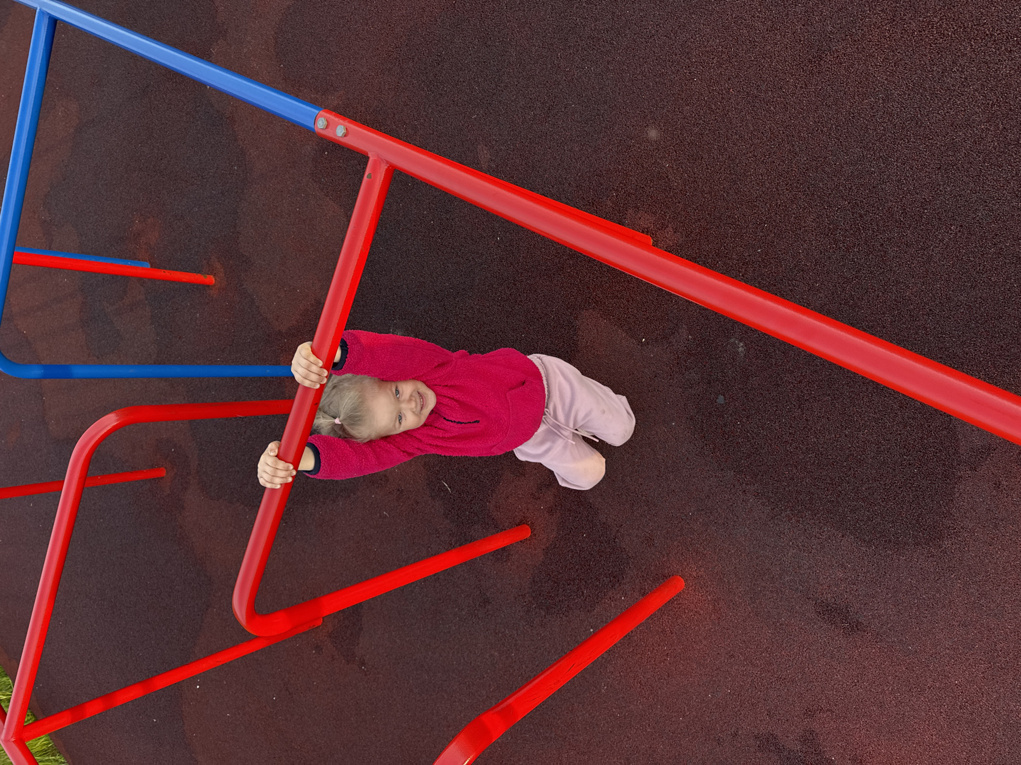 A young blonde child, wearing a fuchsia fleece sweatshirt and light pink sweatpants, hangs from a red horizontal bar on a playground in Горный, Russia. The child's hands grip the bar, and she smiles directly at the camera, with her knees bent and hovering above the dark reddish-brown rubberized safety surface. The metal playground structure consists of prominent red bars in the foreground and midground, with blue horizontal and vertical bars visible in the background to the right. A small patch of green grass is discernible in the upper-left corner of the frame. The image is captured from a high-angle perspective, looking down onto the scene.
