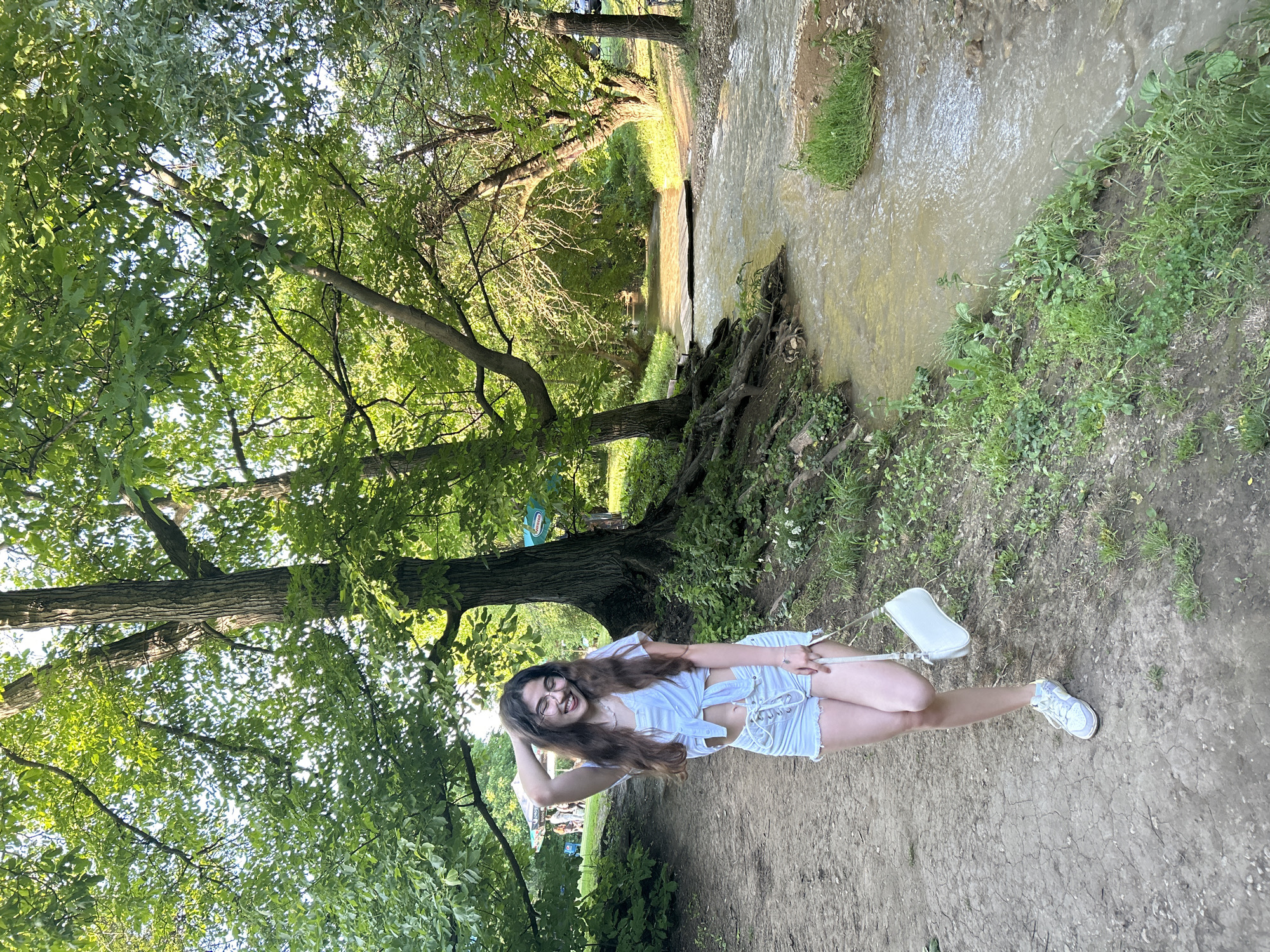 A young woman is standing on a dirt path next to a shallow stream in a wooded area of Hotnitsa, Bulgaria. She is smiling and wearing a white shirt and shorts, with white sneakers and a white purse on her shoulder. The stream is lined with trees and vegetation, and the scene is lit by dappled sunlight. The dirt path is uneven.