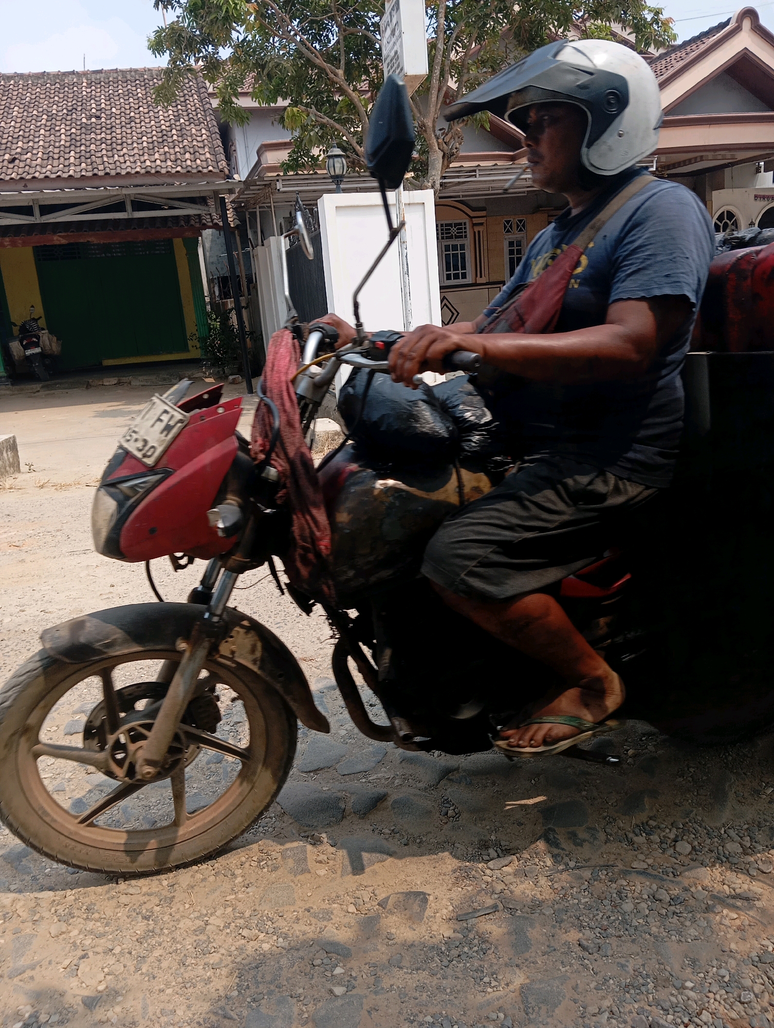 A medium shot captures a person wearing a helmet and a blue t-shirt, riding a red and black motorcycle on a dirt road in Batang Hari Ogan, Indonesia. The rider is holding the handlebars with both hands and has a red bag draped across their chest. A cigarette is visible in the rider's mouth. The motorcycle has a license plate that reads "FW 15-30." In the background, there are houses with tiled roofs and trees. The road is covered in gravel and dirt, suggesting a rural or semi-urban setting. The lighting indicates it is daytime, likely with bright sunlight.