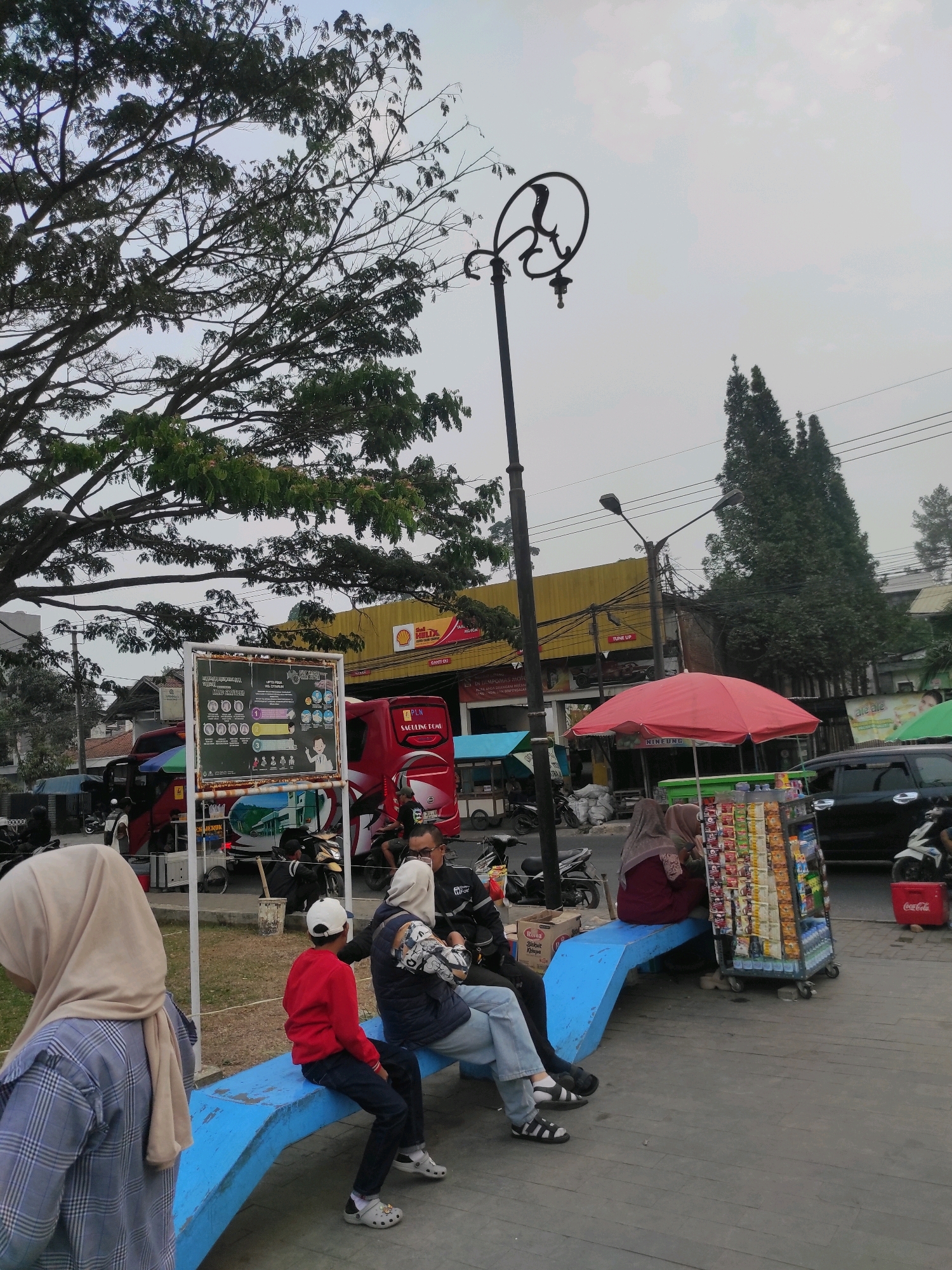 The image depicts an outdoor scene in Padalarang, Indonesia, on a day with overcast skies. A large tree with dense foliage is prominent in the foreground, its branches extending across the top of the frame. To the right, a decorative lamppost with a curved top stands tall. In the middle ground, a group of people are gathered. On the left, a person wearing a blue plaid shirt and a beige hijab is partially visible, facing away from the camera. Beside them, a child in a red jacket and white baseball cap and an adult in a dark jacket and light-colored hijab are seated on a blue bench. Further to the right, two women in hijabs are sitting on the same bench. In front of them is a mobile cart filled with various snacks and drinks, topped with a red umbrella. A black car is parked in the background, and a red Coca-Cola cooler is visible to its right. Behind the bench and cart, several motorcycles are parked, and a red truck with "PLN" (Perusahaan Listrik Negara) branding is visible. A yellow building with "Shell" signage and the text "TUNE UP" can be seen in the distance. A white sign with information and illustrations is also positioned near the truck. The ground is a mix of dirt and wooden planks. The overall atmosphere suggests a public space with commercial activity.