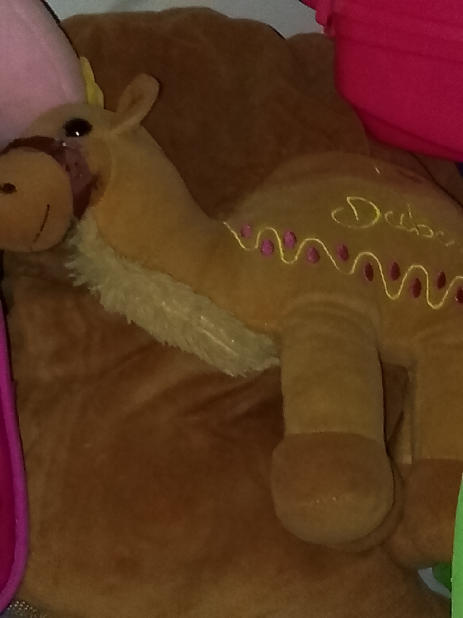 The image features a plush camel toy, predominantly brown, resting on a soft, lighter brown surface, likely another plush item or blanket. The camel toy is positioned on its side, facing towards the left. It has black button eyes and a stitched smile on its snout, which is adorned with a dark reddish-brown bridle-like strap. A lighter, fluffy texture runs along the camel's neck and part of its back, mimicking a mane or hump.
On the camel's side, there are decorative yellow embroidered wavy lines, punctuated by small pink or maroon circular dots. Above this pattern, the word "Dubai" is embroidered in elegant yellow script. The background shows hints of other soft objects or fabrics in various colors, including pink towards the upper left, bright red in the upper right, and a sliver of green at the bottom right.
The scene depicts an inanimate object in an indoor setting, possibly a bedroom or playroom, given the presence of multiple plush items. There is no activity taking place. The lighting is soft and ambient, suggesting an indoor environment without strong natural light. The visible text "Dubai" indicates the theme or origin of the camel toy, likely as a souvenir.