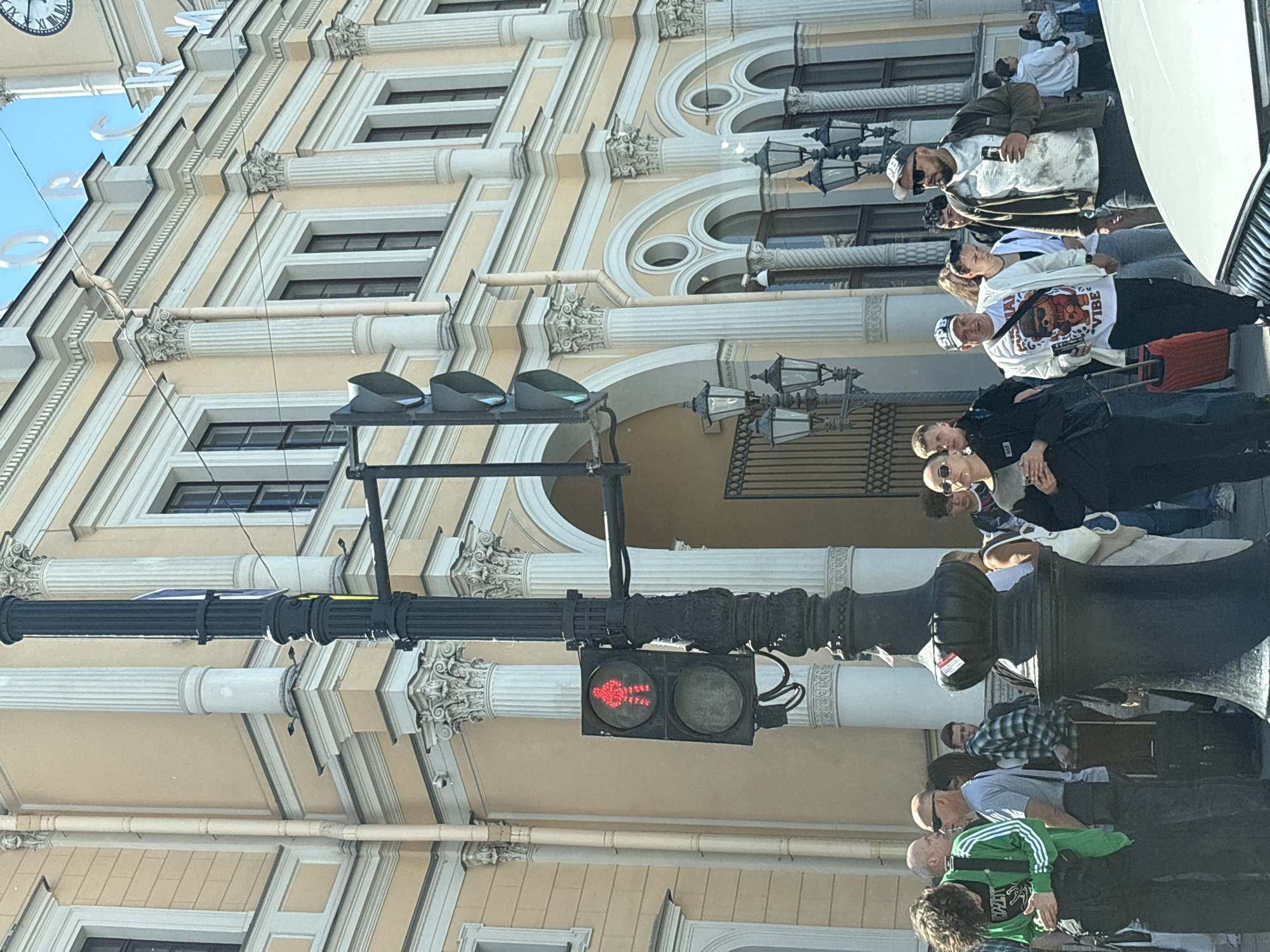 The image depicts a street scene in front of a grand, classical building, likely a train station or other public institution in Saint Petersburg, Russia. The architecture is characterized by tall, fluted columns, arched windows, and ornate detailing, painted in a pale yellow with white trim. A prominent black lamppost stands in the foreground, featuring a traffic signal with a red pedestrian symbol illuminated. Several people are visible, some waiting at the intersection, others walking with luggage. Among them is a person in a green jacket with white stripes, another in a white t-shirt with a cartoon bear graphic, and a man in a brown jacket with sunglasses. The sky is bright and clear. The scene suggests a busy day in a historic urban setting.