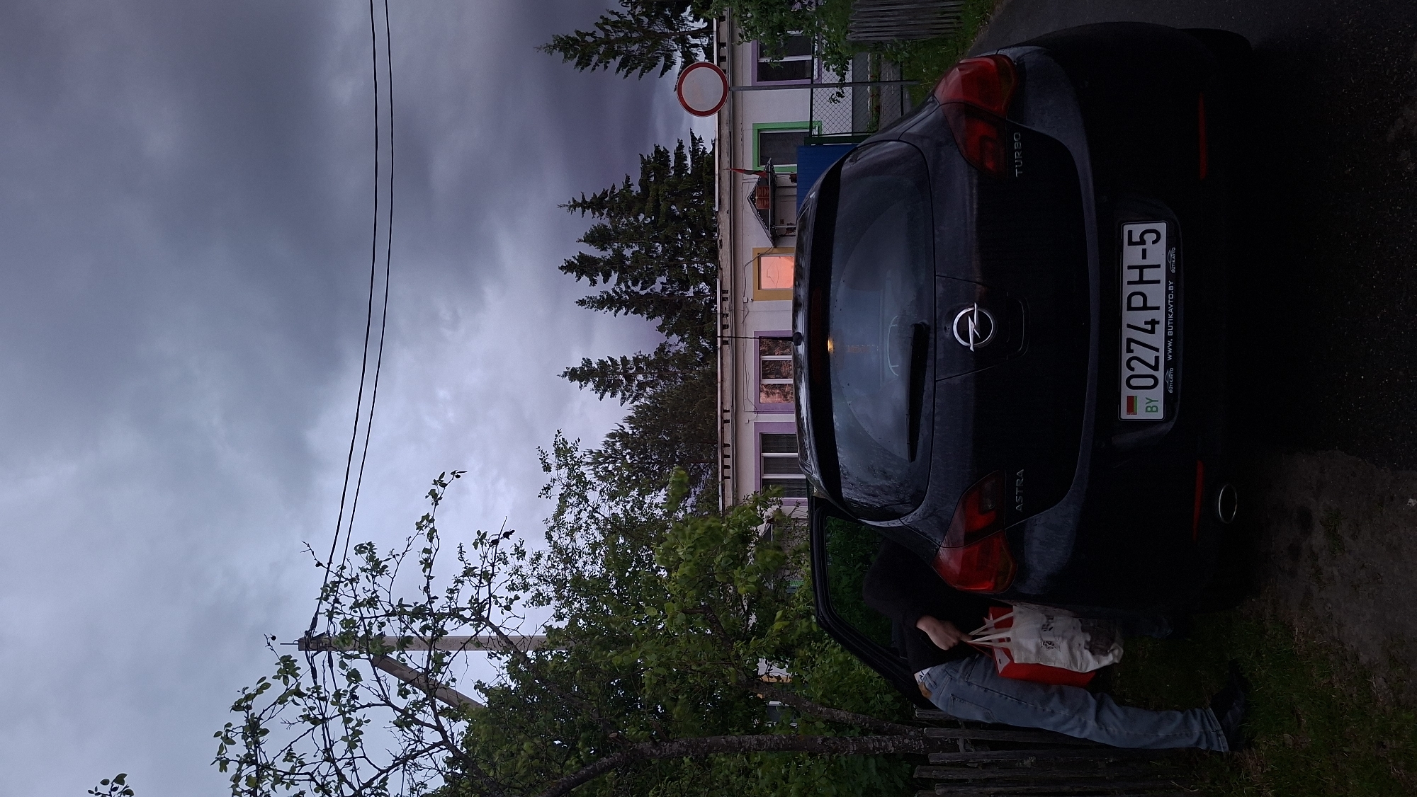 A black Opel Astra, license plate 0274 PH-5 from Belarus, is parked on the side of the road in Забалоцкі сельскі Савет, Belarus. The back of the car is facing the viewer. A person is seated in the driver's seat, with the door open, holding a white and red bag. Overcast sky. A building is behind the car. A No Entry sign is present near the building. Power lines are visible to the left of the building. Trees and shrubbery are in the foreground. A wooden fence is present next to the car.