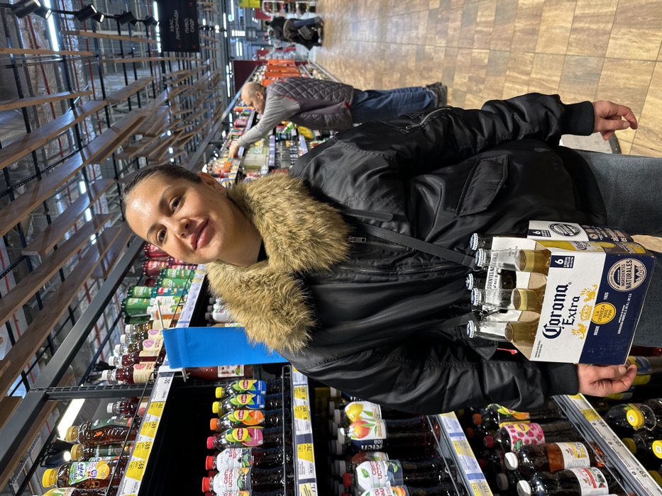 A woman with dark hair pulled back and wearing a black bomber jacket with a light brown fur collar is captured smiling at the camera within a supermarket in Sochi, Russia. She is positioned centrally, carrying a white cardboard six-pack carrier of "Corona Extra" beer bottles slung across her left shoulder via a black strap. The carrier displays brand text including "LA CERVEZA FINA" and "INGREDIENTES NATURALES," and contains six clear glass bottles with blue caps. To the left and immediately behind her, tall supermarket shelves are stocked with various bottled beverages, including Lipton Iced Tea and other juices, displaying price tags in Russian Rubles. The supermarket aisle features a tiled floor with a light brown pattern. In the middle ground, to the right of the primary subject, a bald man in a grey quilted vest and blue jeans is visible, reaching for an item on a shelf. Additional shoppers and grocery carts are present further down the aisle in the background. The ceiling structure is high and industrial, with exposed black beams, modern track lighting, and decorative wooden slats. A red sign in the far background displays Russian text including "КВАС," "ЛИМОНАДЫ," "ПИВО," and "СИДР," confirming a beverage section in a Russian retail establishment.