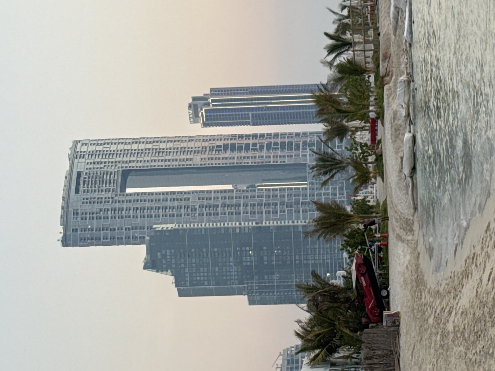 This image captures a waterfront scene in Dubai, United Arab Emirates, likely in the Jumeirah Beach Residence (JBR) area or a similar coastal development known for its modern architecture and beach access. Dominating the background are several towering skyscrapers, characteristic of Dubai's urban landscape. The most prominent structure is a massive, multi-faceted building with a distinctive large open space carved out of its upper section. To its left, a darker, angular skyscraper rises, its facade a mosaic of windows. Further to the right, a slender, illuminated skyscraper stands, displaying vertical lines of blue and white lights, with the word "EMAAR" visible at its top, indicating it's a prominent development.
In the foreground, a sandy beach meets the calm, shallow waters of the Arabian Gulf. A cluster of palm trees and other lush vegetation forms a buffer between the beach and the skyscrapers. Situated on the sand, near the vegetation line, is a striking red sports car, tilted at an angle on a trailer. The car is accompanied by orange traffic cones and a small trailer hitch, suggesting it might be part of an event, display, or possibly a vehicle recovery scenario. The overall atmosphere is serene, with the soft lighting indicating either early morning or late evening. A few sandbags are visible along the shoreline, likely for coastal protection. One person is visible in the distance near the red car, standing near the edge of the vegetation.
