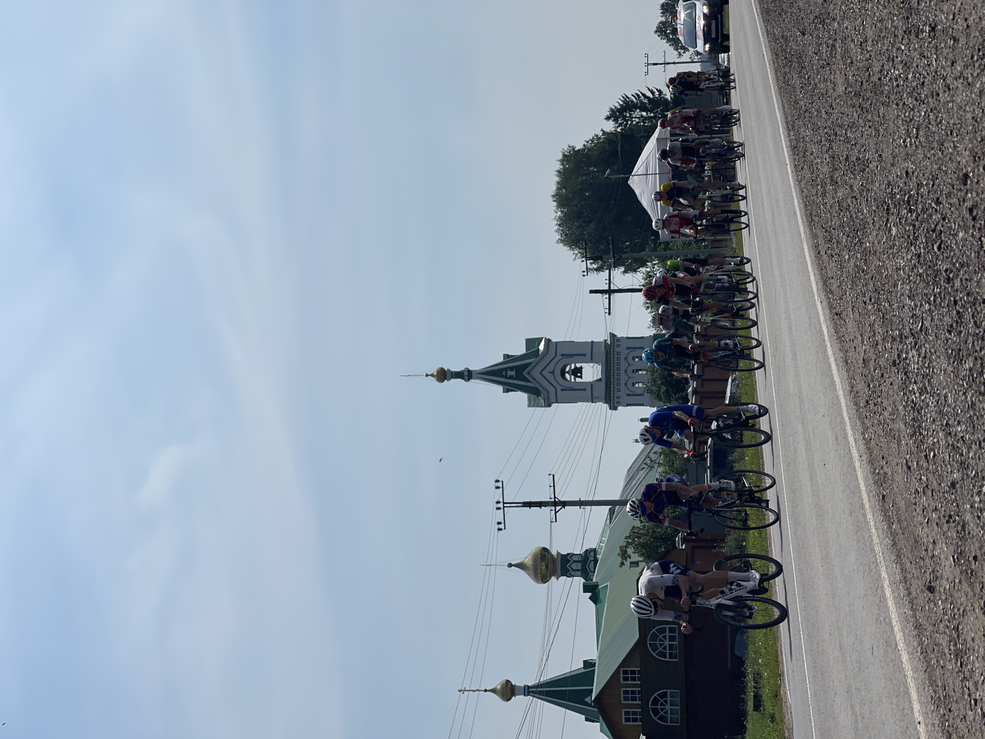 A group of cyclists are participating in a race on a paved road in Perevoznoe, Russia. The cyclists are wearing racing attire and helmets, and are riding road bikes. In the background, there are traditional Russian Orthodox churches with green roofs and golden domes. There are also trees, power lines, and a white canopy tent set up along the side of the road. A white vehicle with a red stripe is parked on the right side of the road. The sky is clear and blue with some wispy clouds.