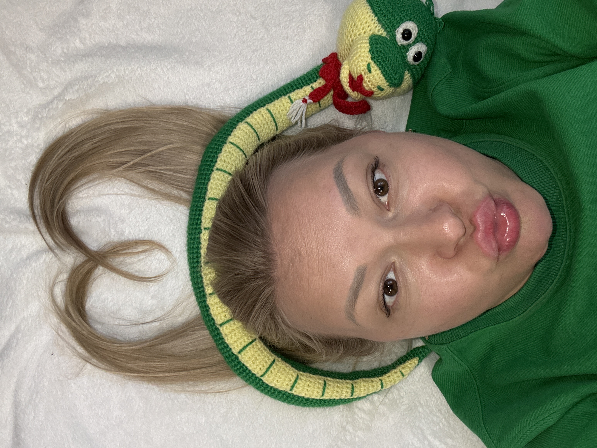 A selfie is presented, showing a Caucasian woman with light brown hair wearing a green sweatshirt and lying on a white, textured surface. Her hair is partially framing her face, with a section styled in a curl. A crocheted, green and yellow snake toy rests on her head. The woman has fair skin, brown eyes, and full lips with what appears to be lip gloss. There is no contextual information to indicate the location is in Izhevsk, Russia.