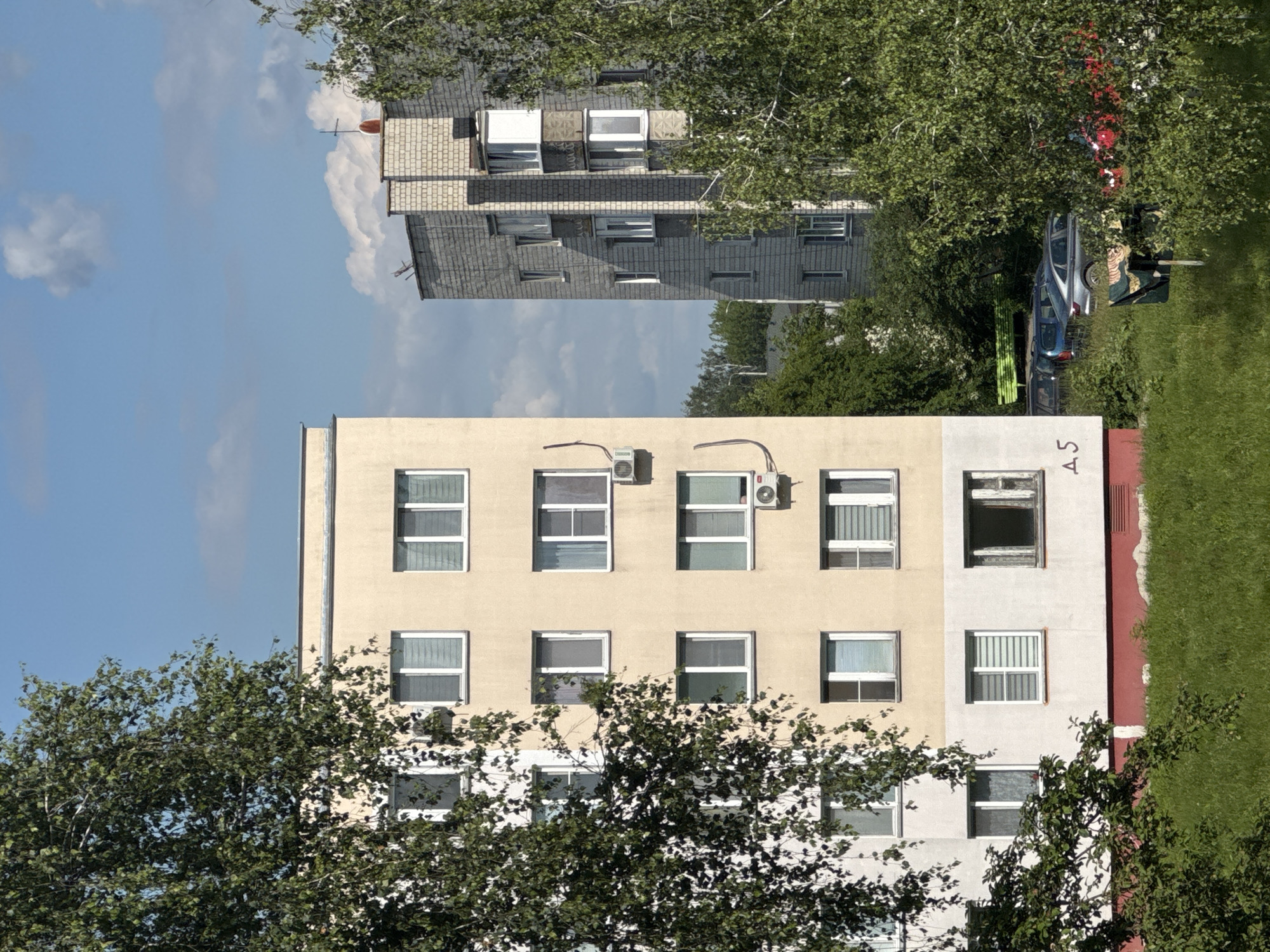 The image shows a view of two apartment buildings in Yarominski Sel'ski Sovet, Belarus, on a sunny day with some clouds. The building in the foreground is a light beige and white multi-story apartment building with numerous windows. A large tree with green foliage partially obscures the left side of this building. The building on the right is a taller, grey brick apartment building with several windows and balconies. The area in front of the buildings is covered in green grass and some trees. Several cars are parked in the background between the buildings. There are no people visible in the image.