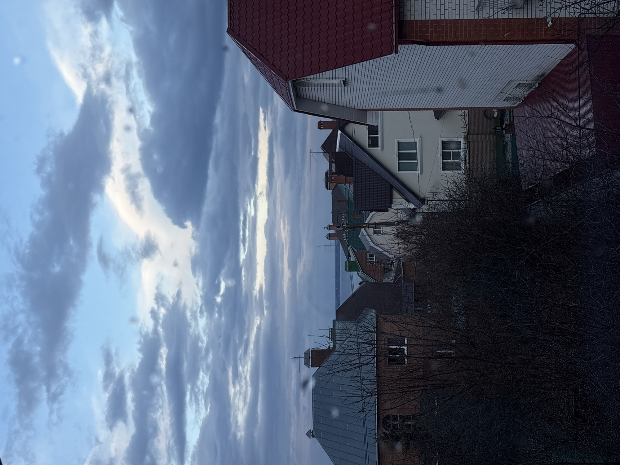 The image, correctly oriented vertically, captures a view of a residential area under a dramatic sky, with a large body of water visible in the distance. The sky, featuring textured clouds ranging from bright white to dark grey against a lighter blue, dominates the upper portion of the scene.
Several houses with varying architectural styles populate the foreground and midground. On the right, a two-story house with a prominent red tiled roof and a mix of white siding and brickwork is clearly visible. Behind it, other houses with dark grey or brown roofs, chimneys, and antennas can be seen, their walls in different shades of light colors, some with visible windows. Bare, dark tree branches and bushes obscure parts of the houses on the right side, suggesting a colder season.
In the middle distance, beyond the houses, a wide, blue-grey expanse of water stretches horizontally, indicating a large river or lake, consistent with the Volga River in Ulyanovsk, Russia.
The lighting suggests either late afternoon/early evening or an overcast day, with diffused light illuminating the scene. Small, blurry specks are scattered across the image, possibly due to dust on the camera lens or water droplets on a window. No people or specific activities are visible, and there is no discernible text. The overall scene conveys a quiet, possibly contemplative atmosphere.
