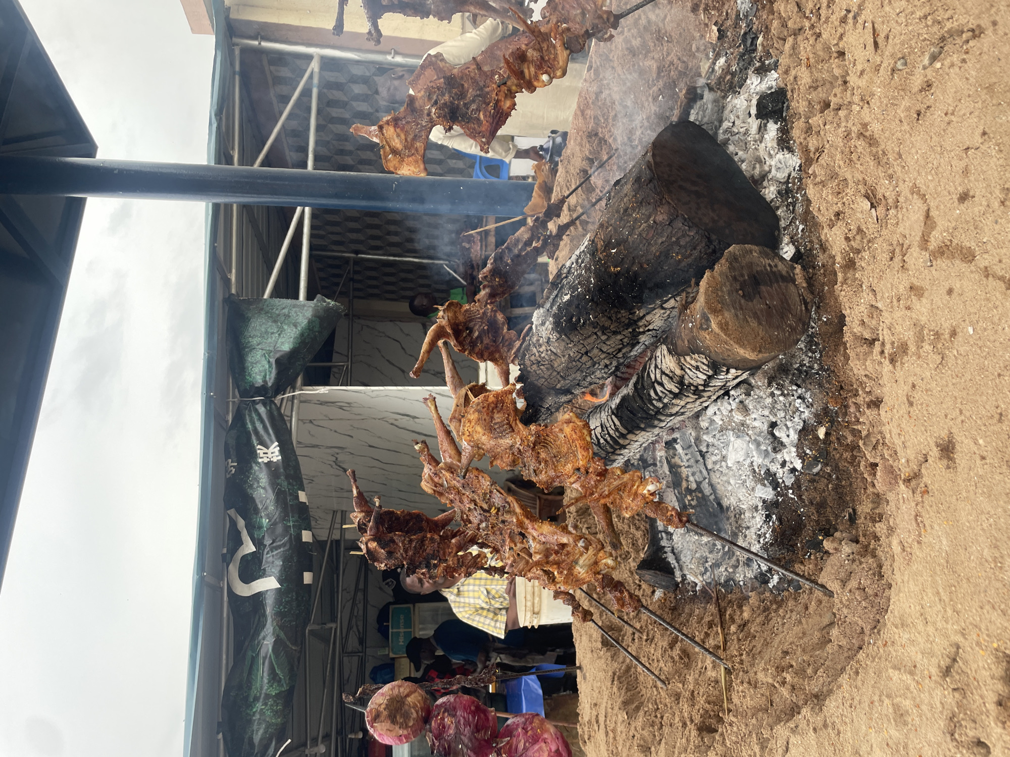This image depicts an outdoor food stall, likely in Abuja, Nigeria, specializing in grilled meat. The foreground is dominated by a pit of hot coals and logs, over which several skewers of what appear to be whole birds or large cuts of meat are being roasted. The meat is browned and appears well-seasoned. The skewers are supported by metal rods inserted into the sand-covered ground.
Behind the grilling area, a man in a yellow and white checkered shirt is visible, standing near a white bucket. To his left, another person in a blue shirt and a black cap is partially visible. In the background, a stall structure with a green and white tarp is set up, suggesting a temporary or semi-permanent vendor location. Signs are visible on the tarp, one with what appears to be Asian characters. Another person in a light-colored shirt is standing to the right, near the grilling area. The overall environment appears to be a dusty, sandy area, possibly a street or an open market, under an overcast sky. There are also some large, round red items, possibly vegetables or decorative elements, hanging on the left side of the image.