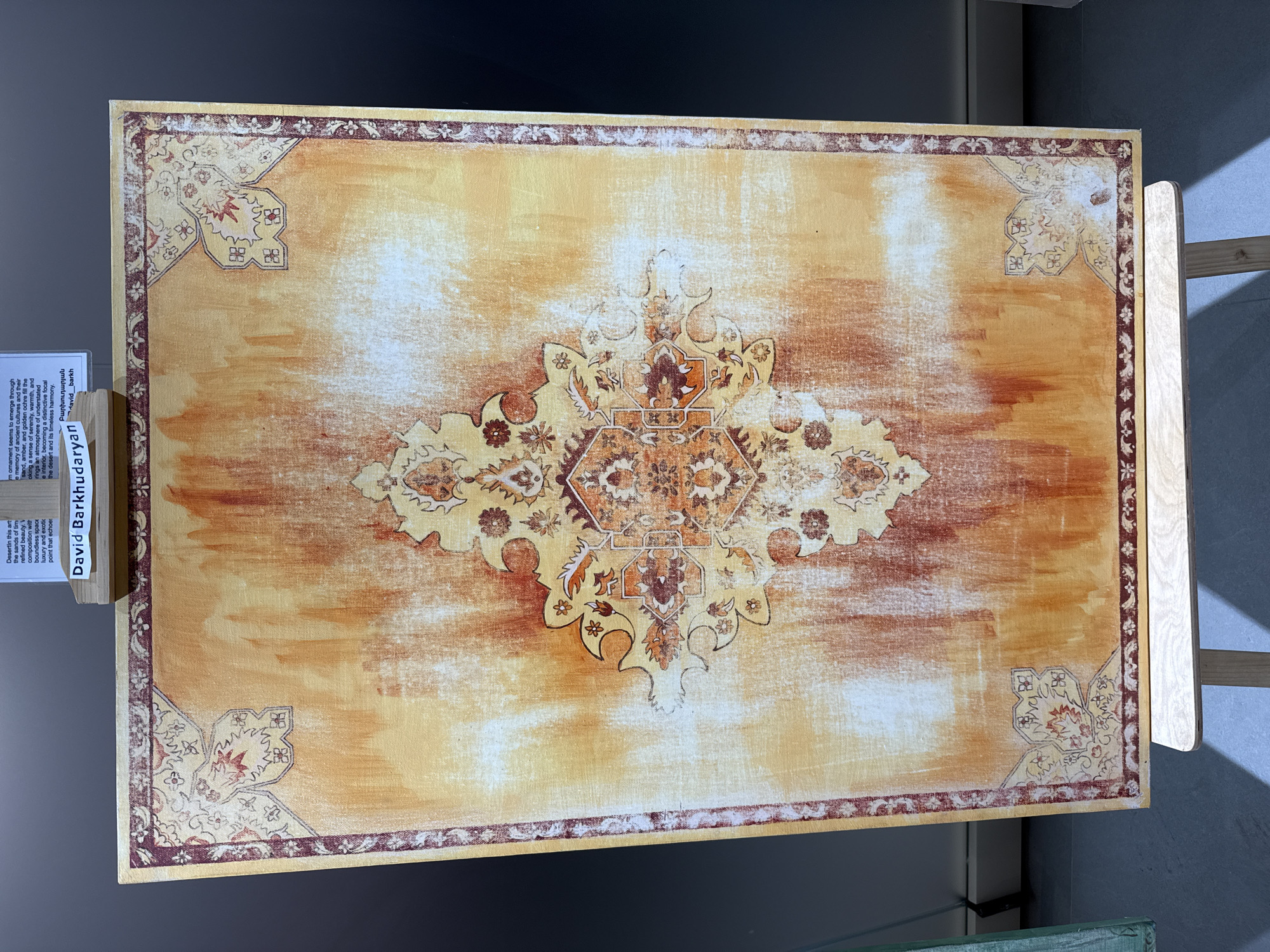 The image shows a framed painting on an easel. The painting is rectangular and has a design reminiscent of a Persian rug, with a central medallion and corner elements. The colors are predominantly in warm tones of orange, yellow, and red, with some white and brown accents. A small plaque with the name "David Barkhudaryan" is visible above the painting. The background is dark, suggesting an indoor setting, possibly an art gallery or exhibition space. Based on the context provided, this artwork is located in Dilijan, Armenia.