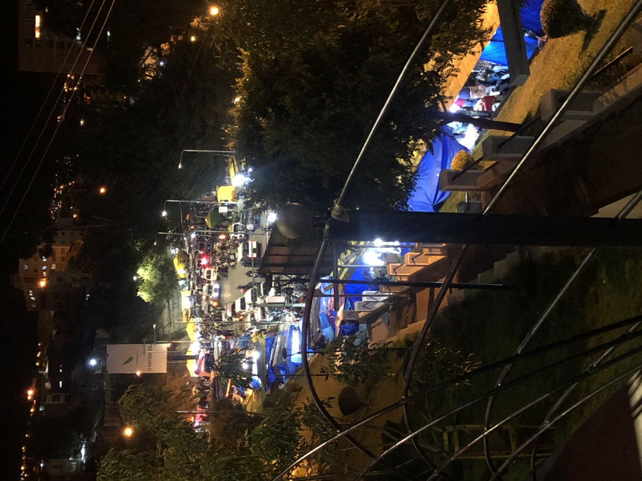 At night, a street crowded with cars and tents illuminated by streetlights presents a busy scene. Blue tents are prominent along the street. Stairs are visible on the right side of the frame. A structure with wires and a pole obscures part of the view. Buildings with illuminated windows line the background.