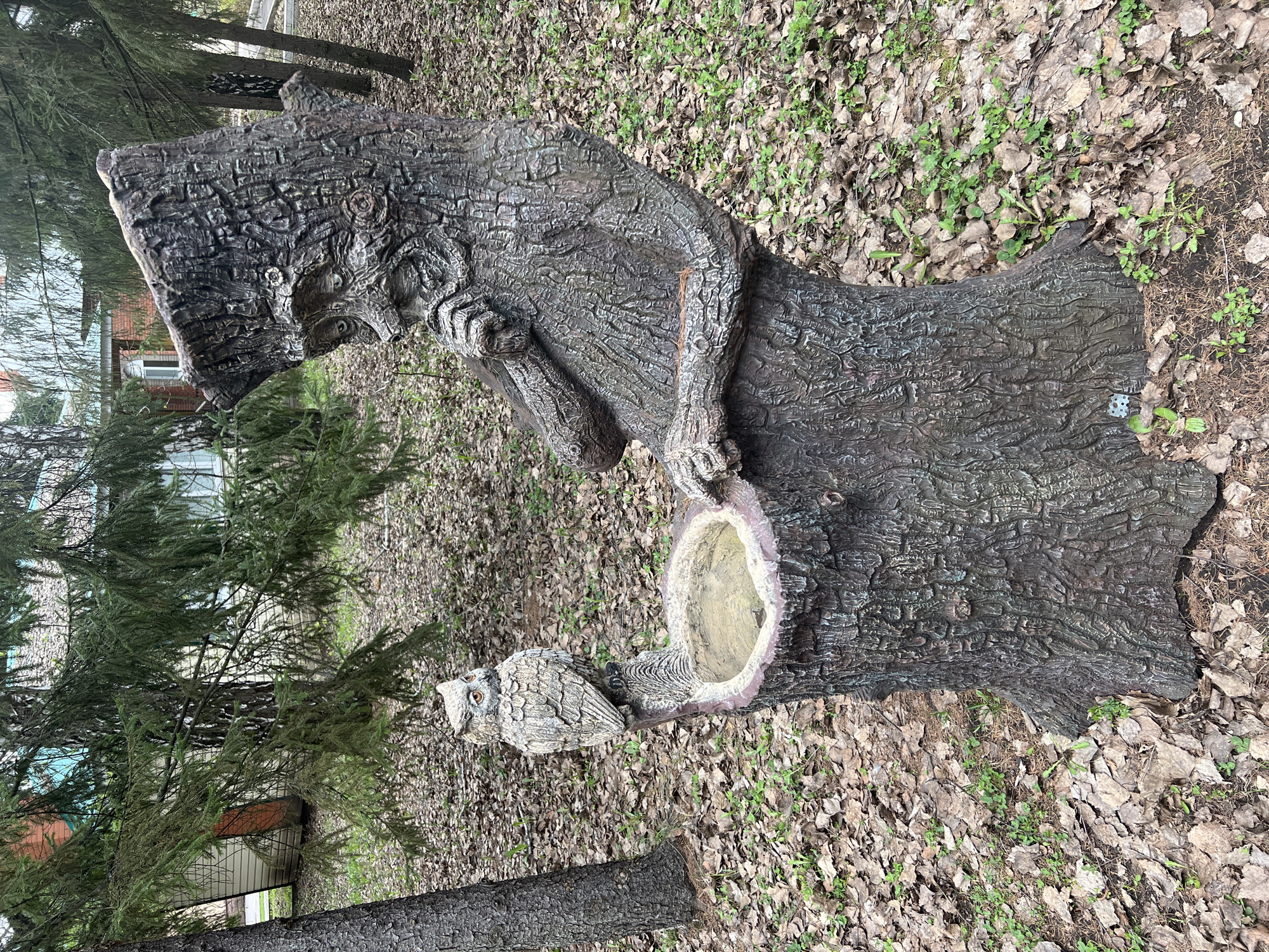 A large, intricately carved wooden sculpture, resembling an old man or forest spirit, stands prominently in an outdoor setting. The sculpture features highly textured surfaces mimicking tree bark, depicting a figure with a long, flowing beard, a hat, and hands placed near a hollowed-out section in its form, which contains some water, possibly serving as a bird bath or planter. An owl sculpture, matching the textured style, is perched on a lower branch-like extension of the main figure.
The ground surrounding the sculptures is covered with a dense layer of dry, fallen leaves, punctuated by small patches of green vegetation. In the background, several coniferous trees, possibly fir or spruce, are visible, creating a wooded atmosphere. Beyond the trees, a light-colored building with a red roof can be partially seen, suggesting the proximity of a residential or institutional structure, consistent with the Sanatoriya Yumatovo Imeni 15-Letiya Bassr location.
The scene is static, with no discernible activity occurring. The lighting is soft and diffuse, indicative of an overcast day or early morning/late afternoon, without strong shadows. The sculptures evoke a sense of folklore and connection to nature. No visible text is present in the image.