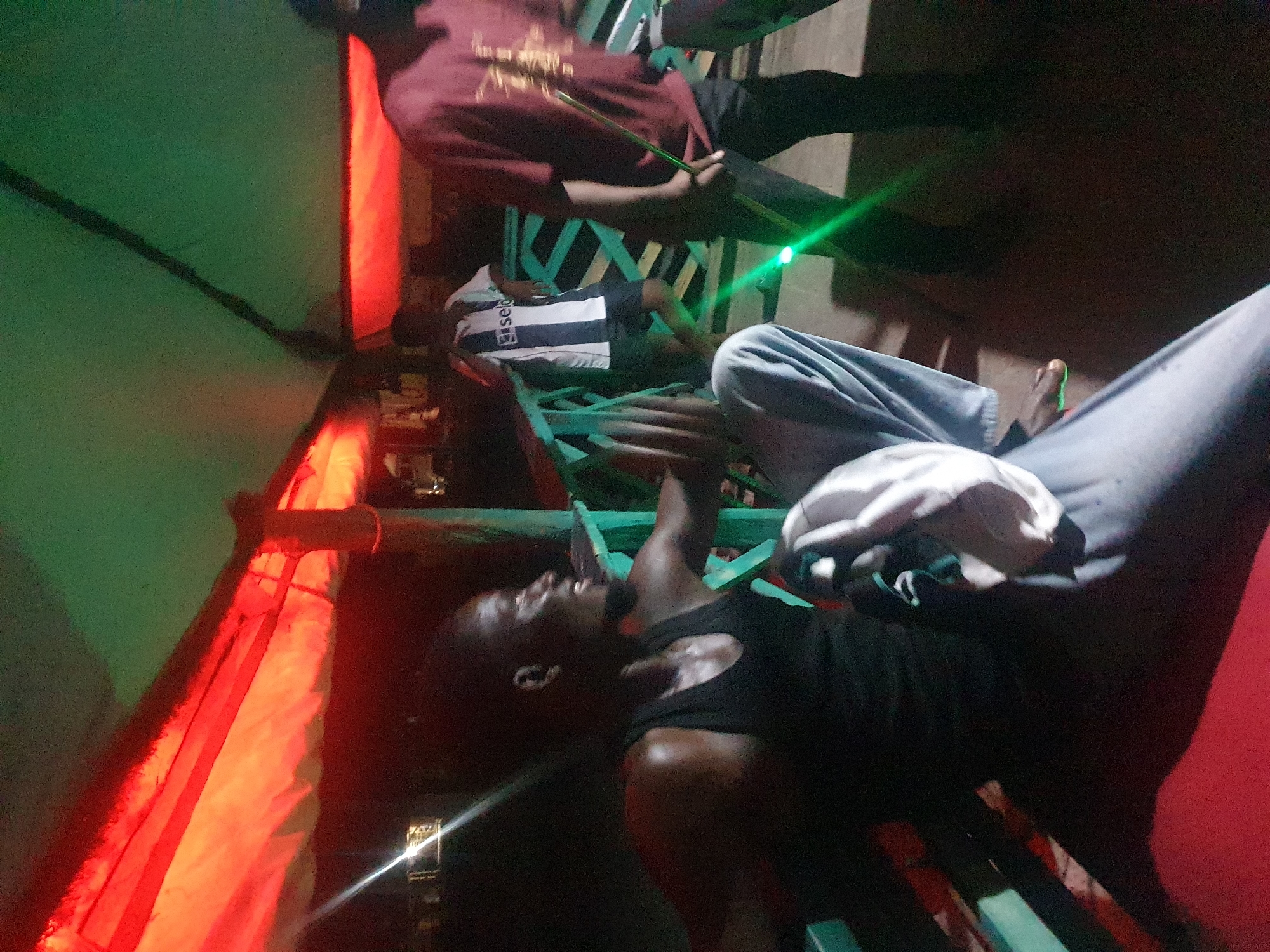 In Lokoja, Nigeria, a group of men gather under a covered structure with red and green lighting. The man in the foreground, wearing a black tank top, is seated and appears to be engaged in conversation or observing something. He has a beard and is looking to his right. To his right, a man in a maroon shirt stands, holding a green glowing stick. In the background, another man is visible wearing a black and white striped jersey. The structure appears to be a covered walkway or seating area, possibly at an outdoor venue or market. The lighting suggests it might be evening or night.