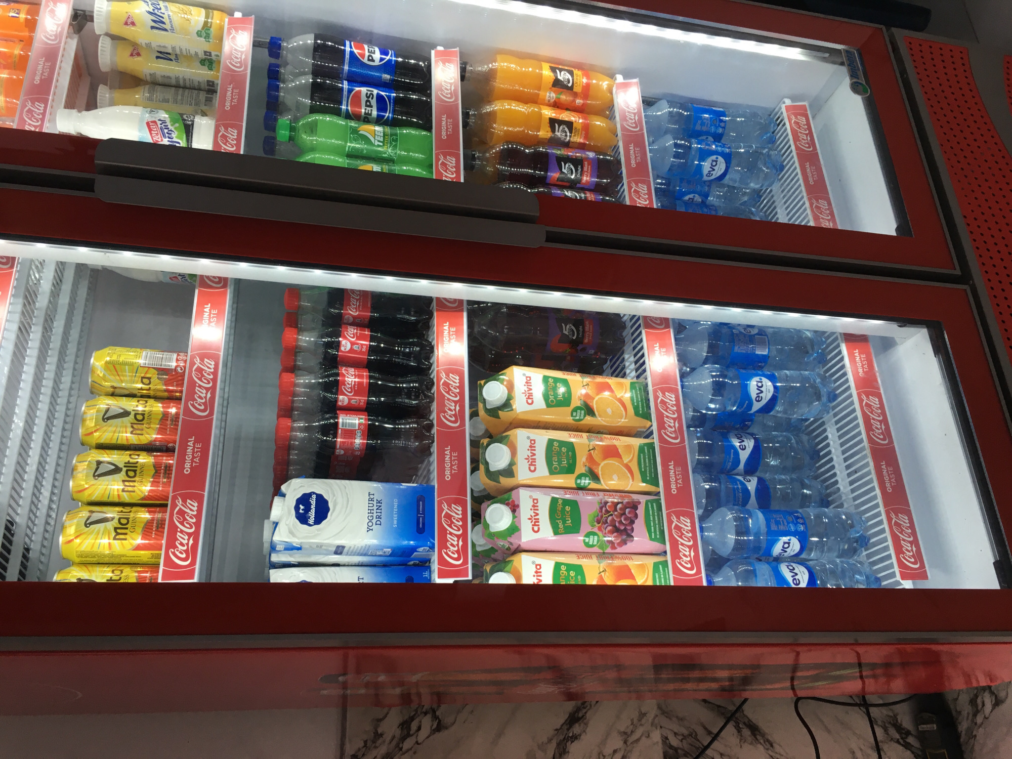 The image shows a refrigerated display case in what appears to be a retail environment in Ado, Nigeria. The case is filled with various beverages, including Malta Guinness, Zetar Yogurt, Coca-Cola, Teem, Pepsi, Chivita juice, and Eva water. Coca-Cola advertising is prominently displayed on the shelf dividers. The case has a red frame and is brightly lit from within. The floor visible in the lower part of the image has a marble-like pattern.