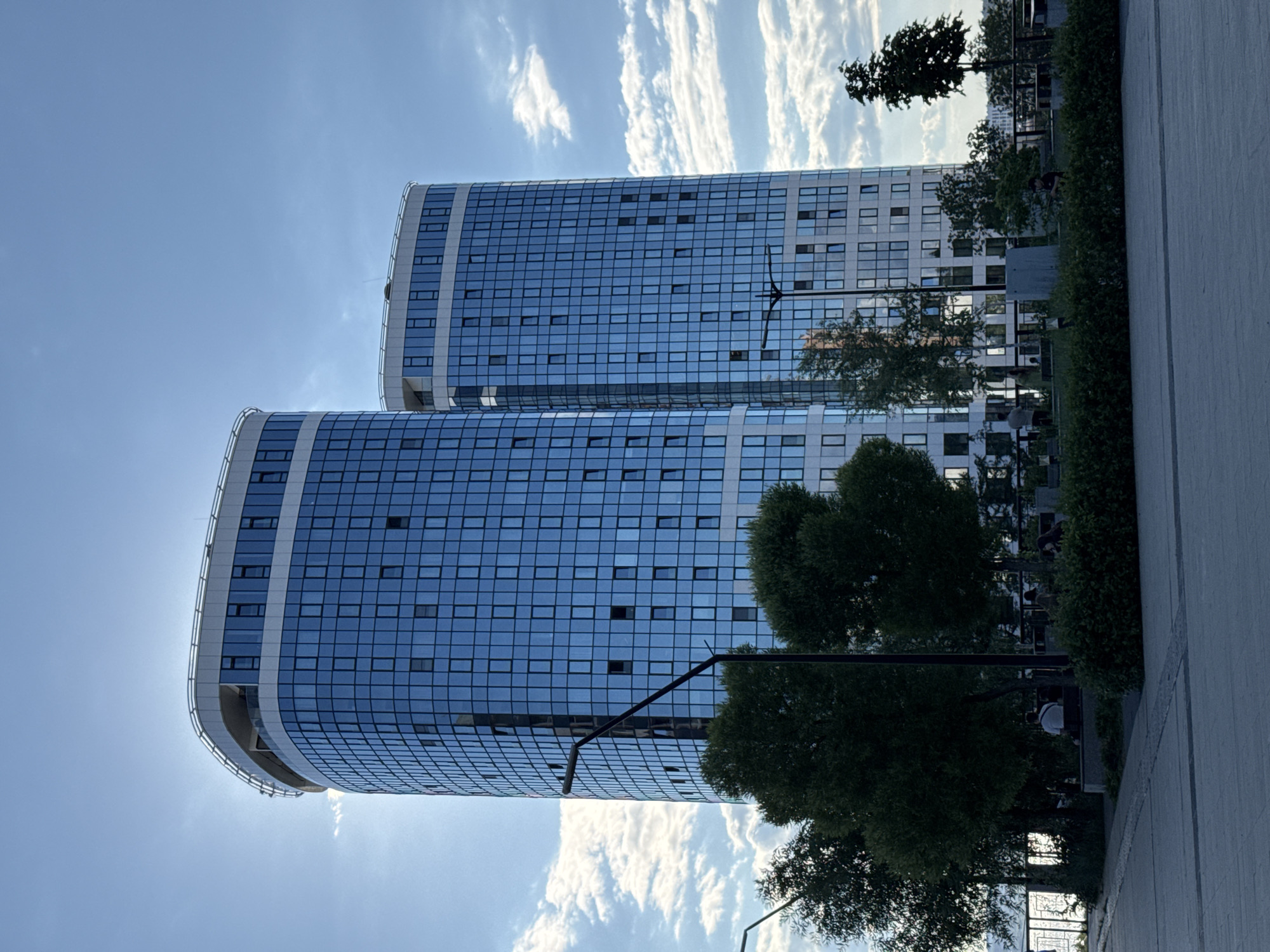 This is a photograph of the "Pearl of the Kama" residential complex in Izhevsk, Russia. The image features two prominent, modern skyscrapers with distinctive curved facades covered in blue glass. The buildings are tall and sleek, with a white trim along the top. The sky above is partly cloudy with a bright, diffused sun peeking from behind one of the buildings. In the foreground, a well-maintained park area is visible with lush green trees and manicured bushes. Several people are seated on benches in the park, enjoying the outdoor space. A paved walkway or plaza runs in front of the park, leading towards the viewer. The overall scene depicts a contemporary urban environment with a blend of modern architecture and green recreational spaces.