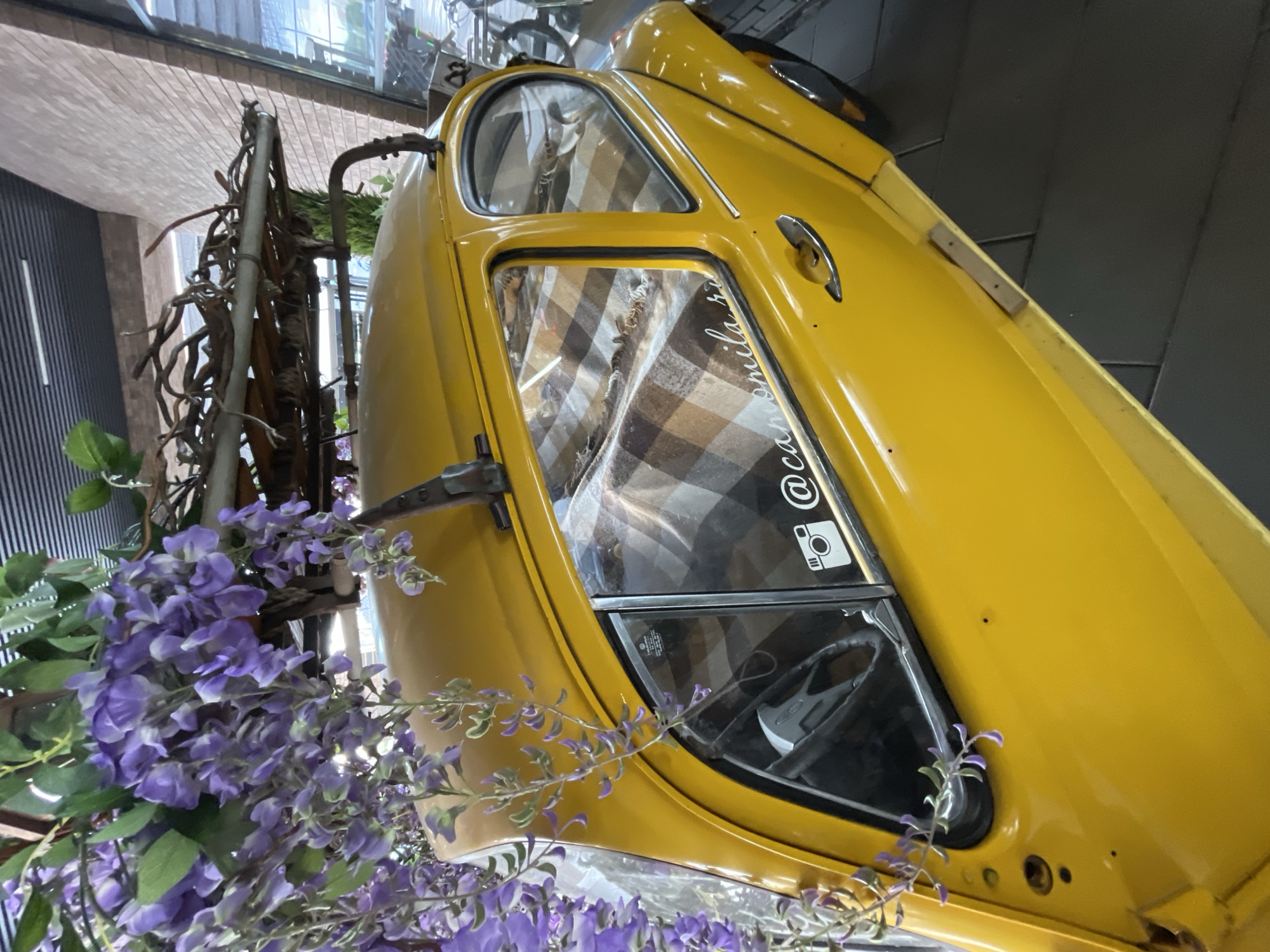 The image shows a yellow vintage Volkswagen Beetle parked outdoors, possibly in a city. The car is adorned with purple wisteria flowers cascading from a wooden rack on its roof. Through the windows, a checkered blanket is visible on the seats. A sticker with a camera icon and text is on the rear passenger window. The background reveals a brick wall and a modern building with a glass facade, suggesting an urban setting. The ground is paved with dark tiles. There are no people in the image.