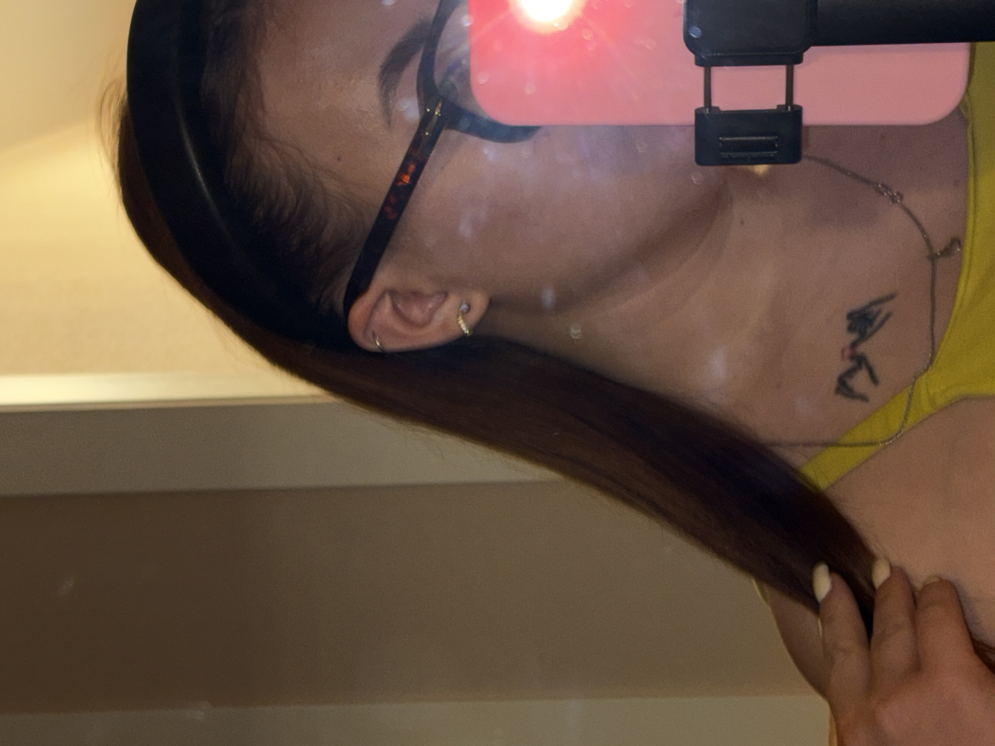 A person's profile is captured in a mirror selfie. The individual wears glasses with tortoise-shell frames, a black headband, and a yellow top. They have a tattoo on their upper chest and multiple earrings. Part of a phone with a pink case and a black phone holder attachment is visible in the foreground. The background is a beige wall. There is no information to suggest the location is in Krasnodar, Russia.