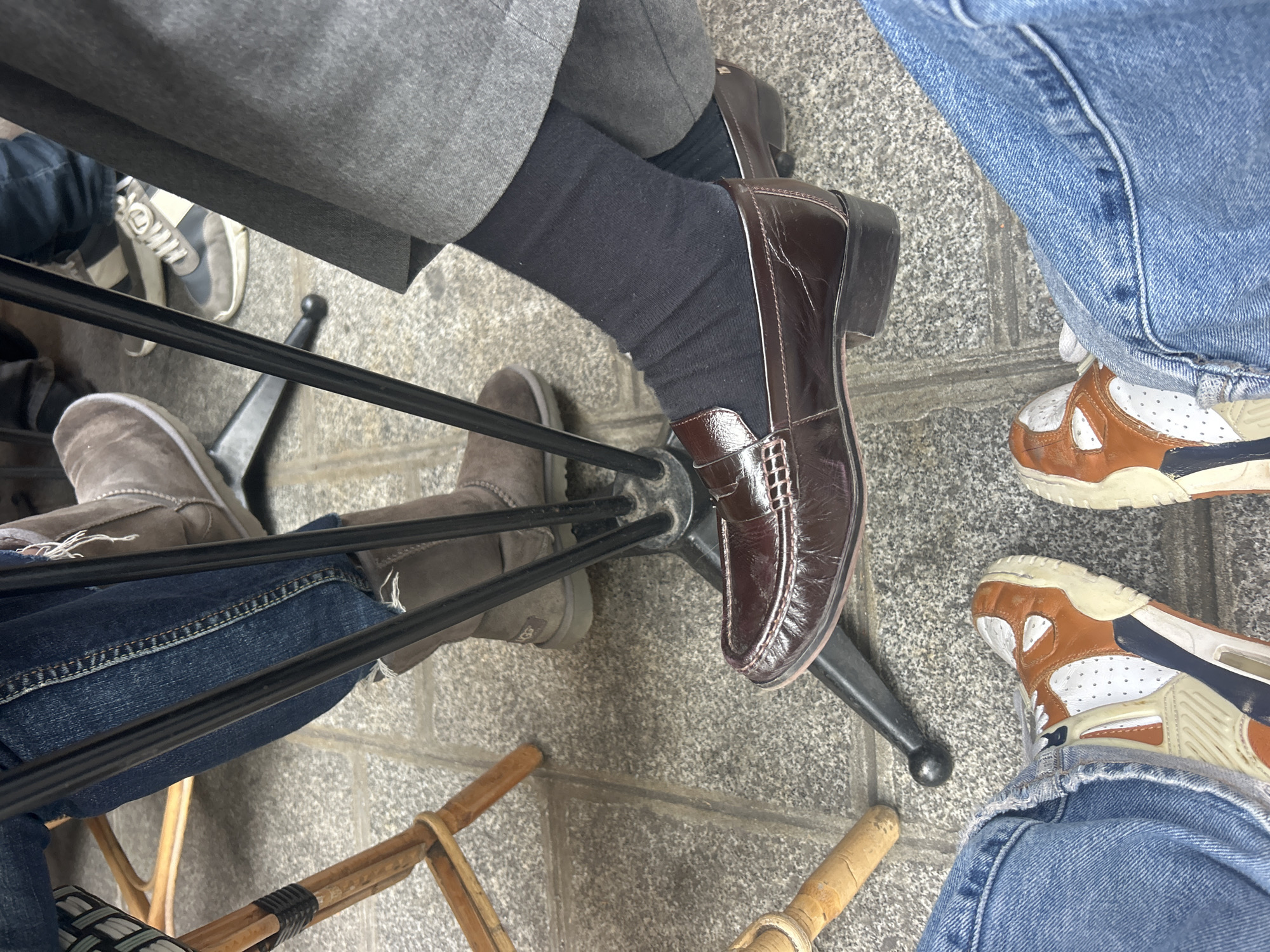 Captured from an elevated perspective, the image depicts the lower portions of three individuals seated in an urban setting. The setting appears to be a public space, possibly a cafe or outdoor dining area, suggested by the stone-tiled ground and an assortment of chair types including metal and wood-based designs.
The individual on the left wears blue jeans and casual taupe-colored boots. In the middle, a person is dressed in a gray suit and wears formal brown leather loafers over dark socks. To the right, another individual is seen in blue jeans and sport shoes with a tan, white, and blue color scheme. The arrangement of their legs and the proximity of the chair legs indicate they are likely seated around a table.