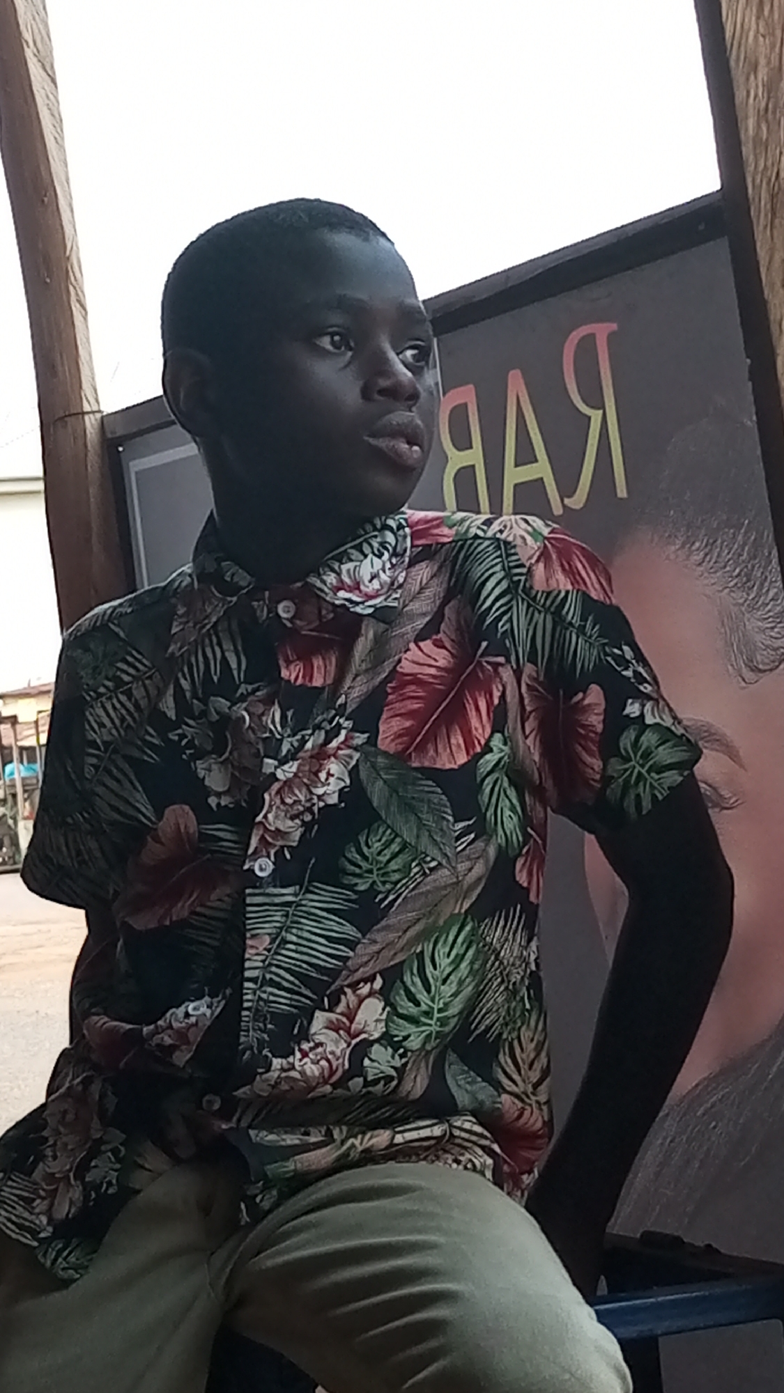 In Sabon Tasha, Nigeria, a young, dark-skinned person wearing a colorful floral-patterned shirt sits on a chair. The person is looking off-camera to the right. Behind the person is a sign with the word "BAR" visible, suggesting a business establishment. The surrounding environment is shaded, with elements of an urban street visible in the background. The person is wearing khaki-colored trousers.