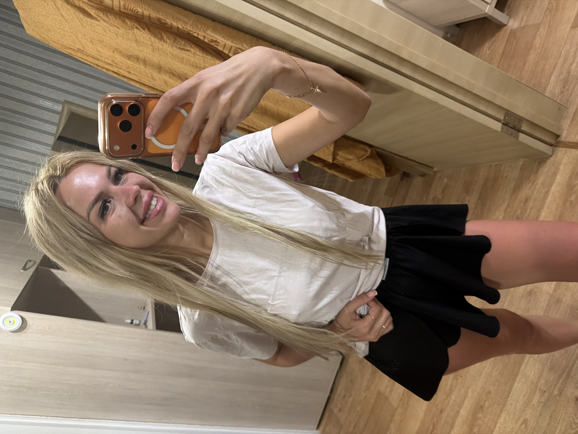 A blonde woman is taking a mirror selfie in what appears to be a bedroom or dressing room. She is wearing a white t-shirt and a black skirt, and she has a phone in her hand. The room has wooden floors and light-colored cabinets. The woman is smiling and looking at the camera. There is a yellow curtain in the background. The image does not provide any specific details or context that would allow for an accurate determination of the location within the city of Kyiv, Ukraine.