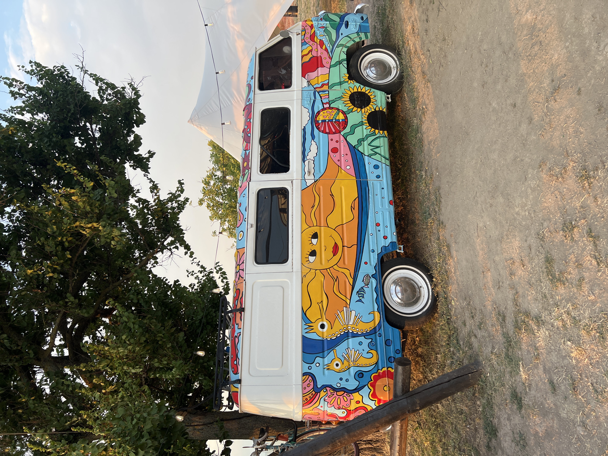 A vibrantly painted vintage van with a colorful mural depicting a sun, seahorses, fish, flowers, and abstract wavy patterns is parked on a dirt path. The van has white upper sections and a white roof rack. A large tree with green leaves overhangs the left side of the van, and string lights are visible hanging from its branches. In the background, a large white tent is partially visible, along with other trees and a bright sky with scattered clouds. A wooden post stands in the foreground to the left of the van. The setting appears to be an outdoor event or campsite, possibly near the city of Yeysk, Russia. There are no people visible in the image.