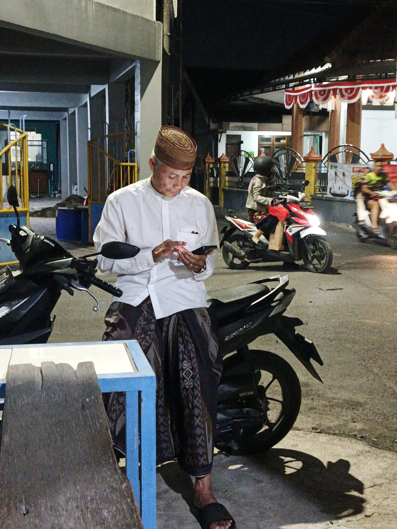 This is a full shot image of a man in Ketegan, Indonesia, at night. The man is wearing a white long-sleeved shirt, a patterned sarong, and a woven cap. He is standing next to a black motorcycle and looking down at his phone. The background shows a building with red and white banners, a street with other motorcycles and people, and trees. The lighting suggests it is evening or nighttime.