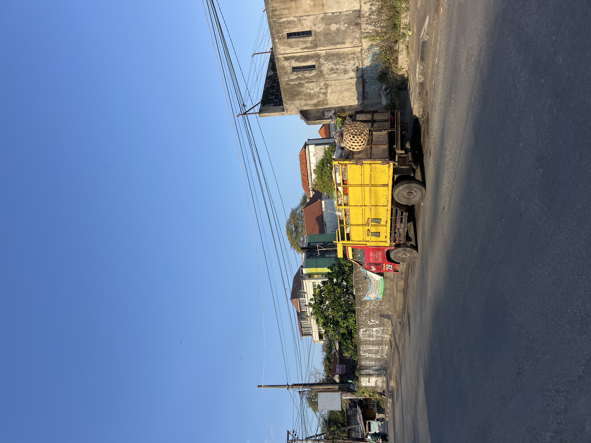 An eye-level, medium-shot captures a bright yellow truck parked on the side of a paved road in Denpasar, Indonesia. The truck, which appears to be a utility vehicle, is loaded with various items, including what look like large woven baskets and bundled materials. A red cab with the number 79 prominently displayed on the side is visible. The truck is positioned next to a concrete wall, which is covered in graffiti and weathered. Beyond the wall, several buildings are visible, including a multi-story structure with a light-colored facade and an orange-tiled roof, and another building with a green accent. A power pole with numerous wires stretches across the frame, indicating an urban or semi-urban setting. The sky above is a clear, bright blue. To the left of the truck, a portion of another vehicle, possibly a scooter, is seen on the road. The overall scene suggests a sunny day, likely in the morning or afternoon, given the shadows cast on the road.