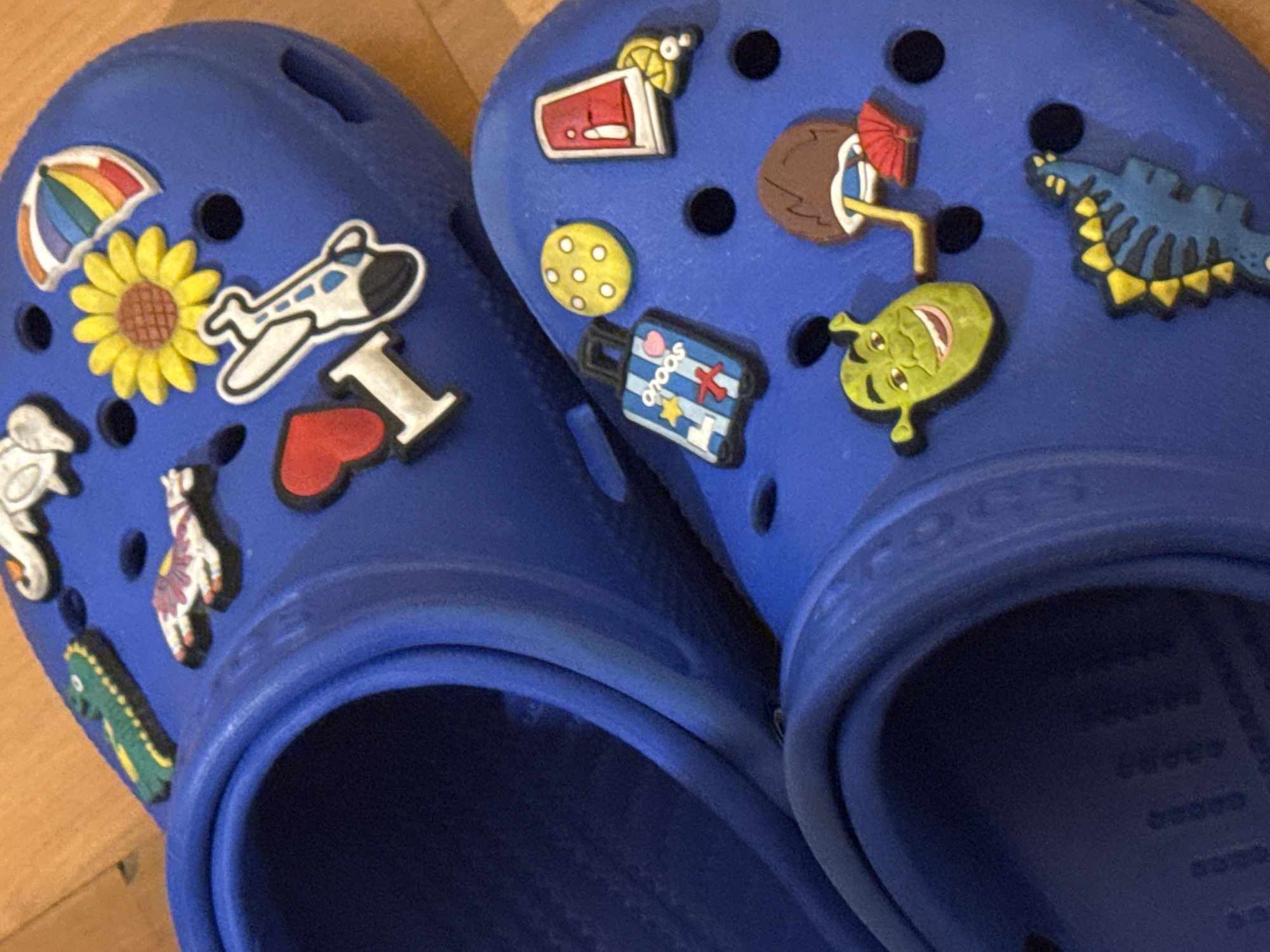 Two blue Crocs shoes are displayed. The left shoe has a rainbow umbrella, a sunflower, an airplane, a red heart, an "I" letter, a seahorse, a skeleton hand, and a small green alligator charm. The right shoe is adorned with a red cocktail charm, a cookie charm, a travel bag charm, a coconut drink charm, a Shrek charm, and a blue dinosaur charm. The shoes are sitting on a light wooden surface. The location is not discernible from the image.