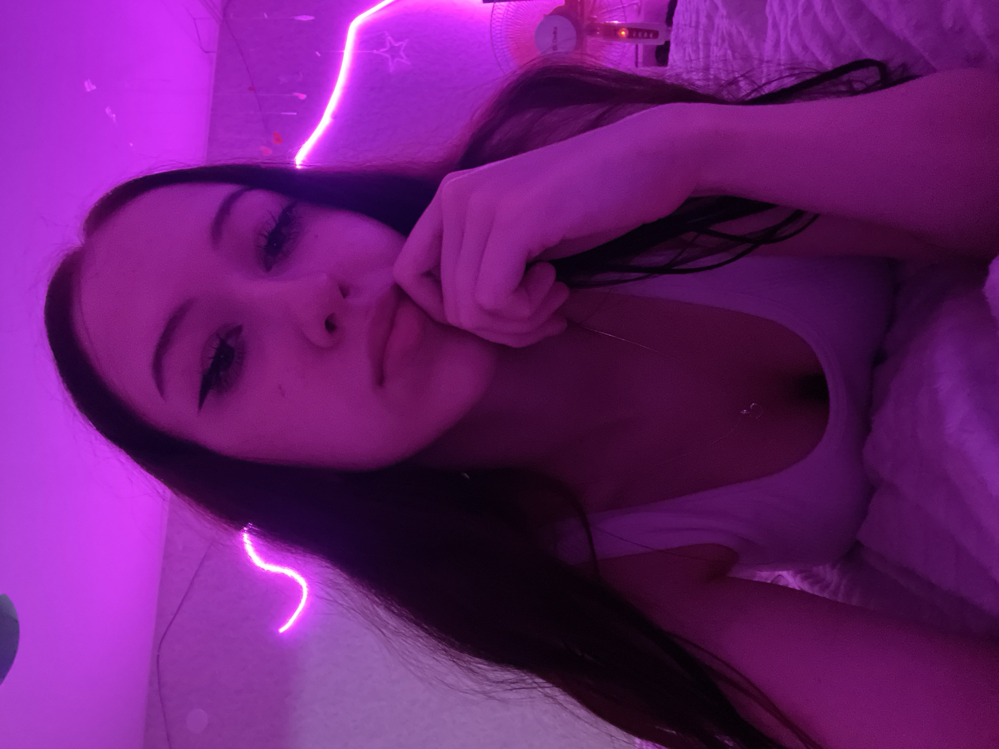 The image depicts a close-up, selfie-style shot of a young woman with long, dark hair, lying on a bed. She is illuminated by pink neon lighting, which casts a strong purple hue over the scene. The woman is wearing a white tank top and has her hand resting near her chin, with a pensive expression. The background features the pink neon lights outlining a wavy shape on the wall, and a small fan is visible in the upper right corner. There is no discernible information about the location in Votkinsk, Russia.