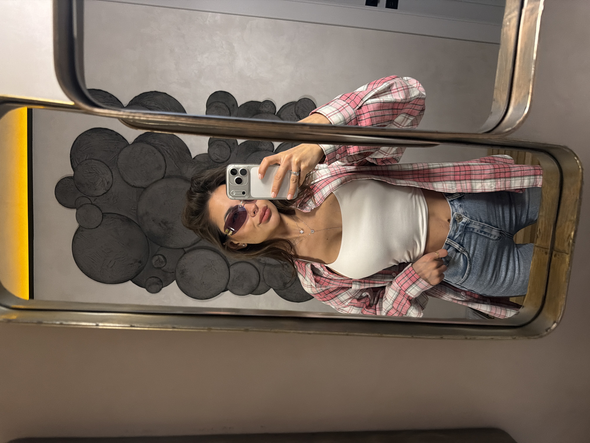 A woman stands in front of a mirror, taking a selfie. The mirror's frame is gold. She is wearing sunglasses, a white tank top, a pink and white plaid shirt, and blue jeans. Behind her, the wall is decorated with dark gray circles. The location appears to be indoors, possibly in a cafe or establishment in Krasnoyarsk, Russia, given the context of the query.