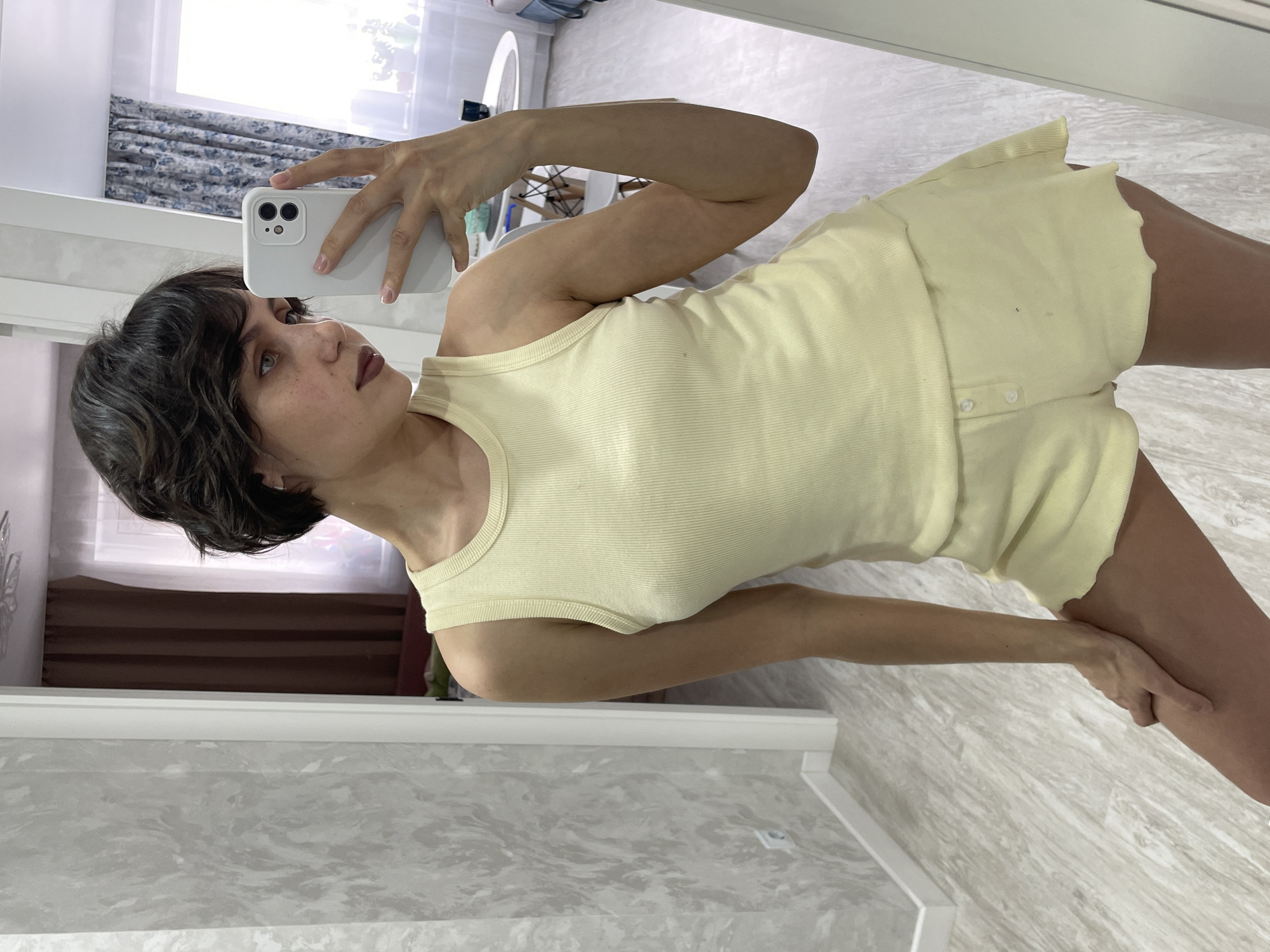 A woman stands indoors, taking a selfie in a mirror with her iPhone. She is dressed in a yellow ribbed tank top and matching shorts. The location appears to be a room with marble flooring and white trim. A window with patterned curtains is visible in the background. The scene does not appear to be in Izhevsk, Russia, as there are no contextual clues to support this location.
