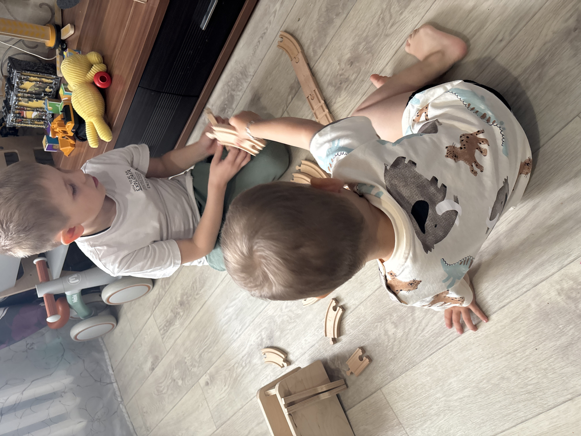 An overhead, slightly angled shot captures two young children engaged in play on a light wood-patterned floor indoors. The child on the left, facing towards the right, has light brown hair and wears a white t-shirt with visible text including "TO BE ME EXPLORER" and "ENJOY". He is seated with green bottoms and is holding several pieces of wooden toy train tracks.
To his right, another child, seen from the back and in profile, also has light brown hair. This child is wearing a cream-colored top patterned with various animal figures, such as bears and leopards, and is barefoot. He is leaning forward, focused on several wooden train track pieces scattered on the floor directly in front of him, along with a small wooden stand or structure. Both children appear to be actively involved in assembling or arranging the toy train set.
In the background, near a dark brown piece of furniture, various other toys are visible, including a yellow stuffed toy, a red ring, colorful building blocks, and a yellow toy truck. A grey and orange balance bike with the word "mini" printed on its frame is partially visible to the far left. The scene suggests a calm, focused playtime, likely during the daytime, given the soft indoor lighting.